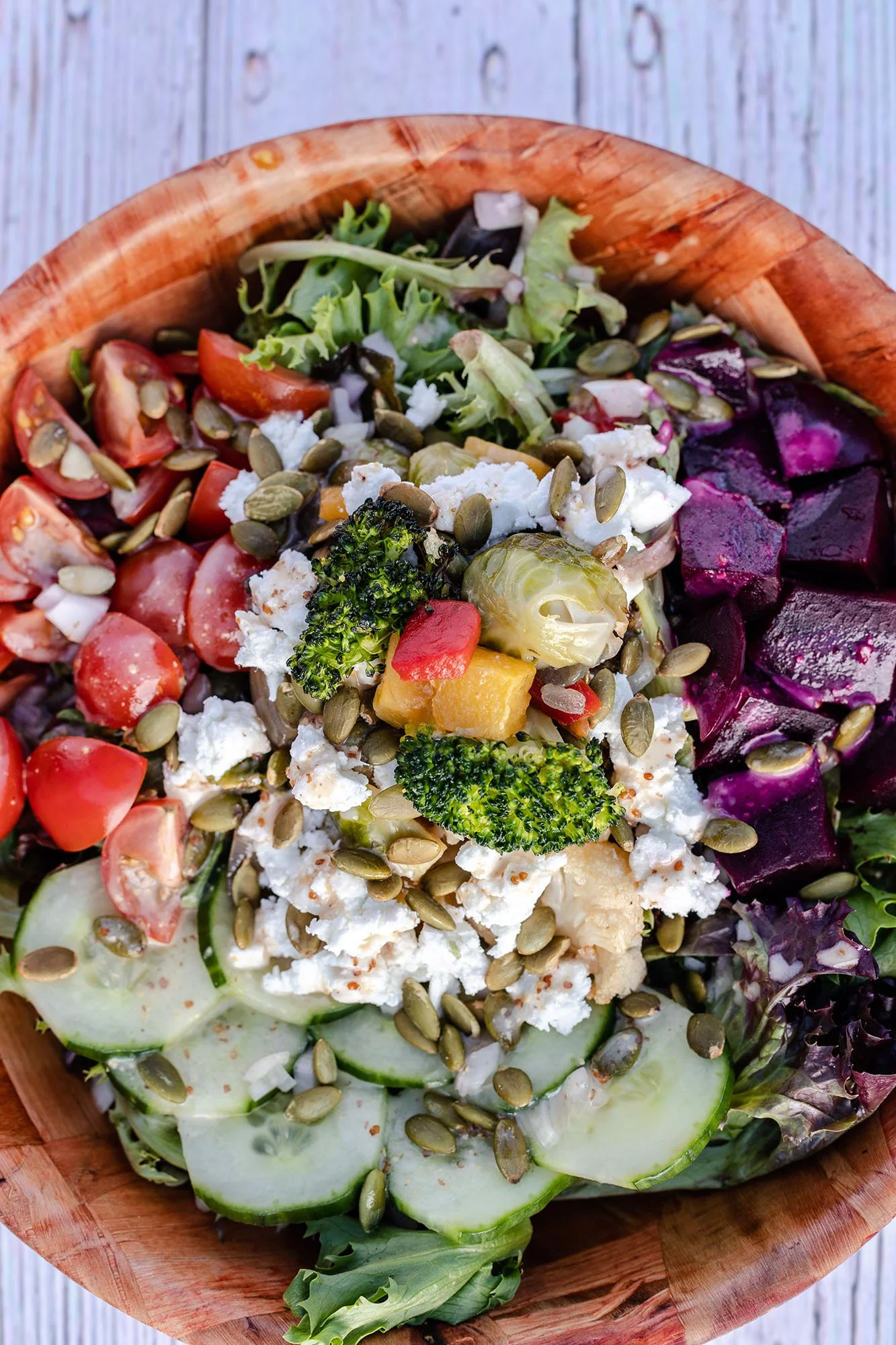 A colorful salad in a wooden bowl with cucumber, cherry tomatoes, broccoli, roasted parsnips, mixed greens, feta cheese, sunflower seeds, and shredded purple beets.
