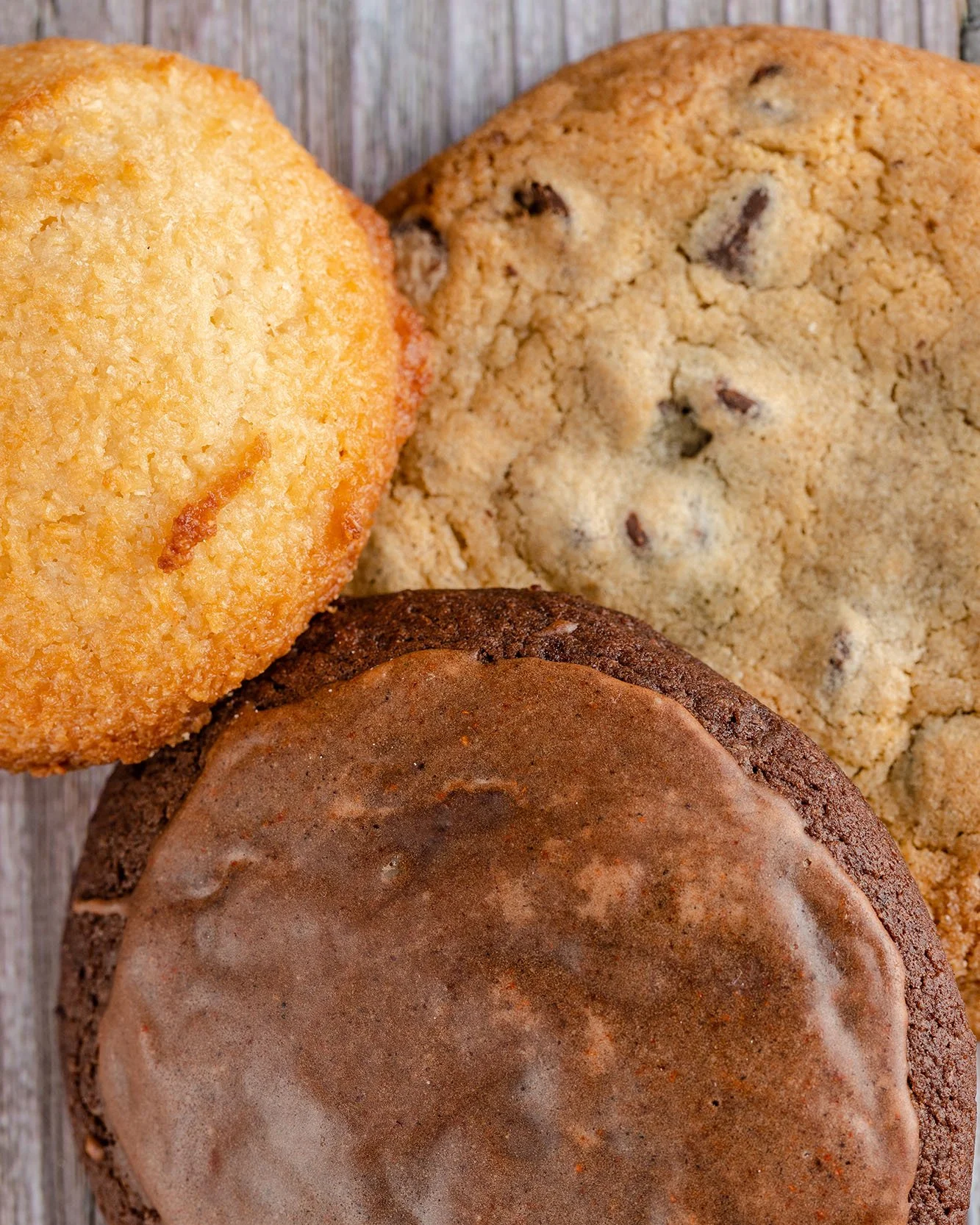 Close-up of three different cookies on a wooden surface, including a sugar cookie, chocolate chip cookie, and a glazed cookie.