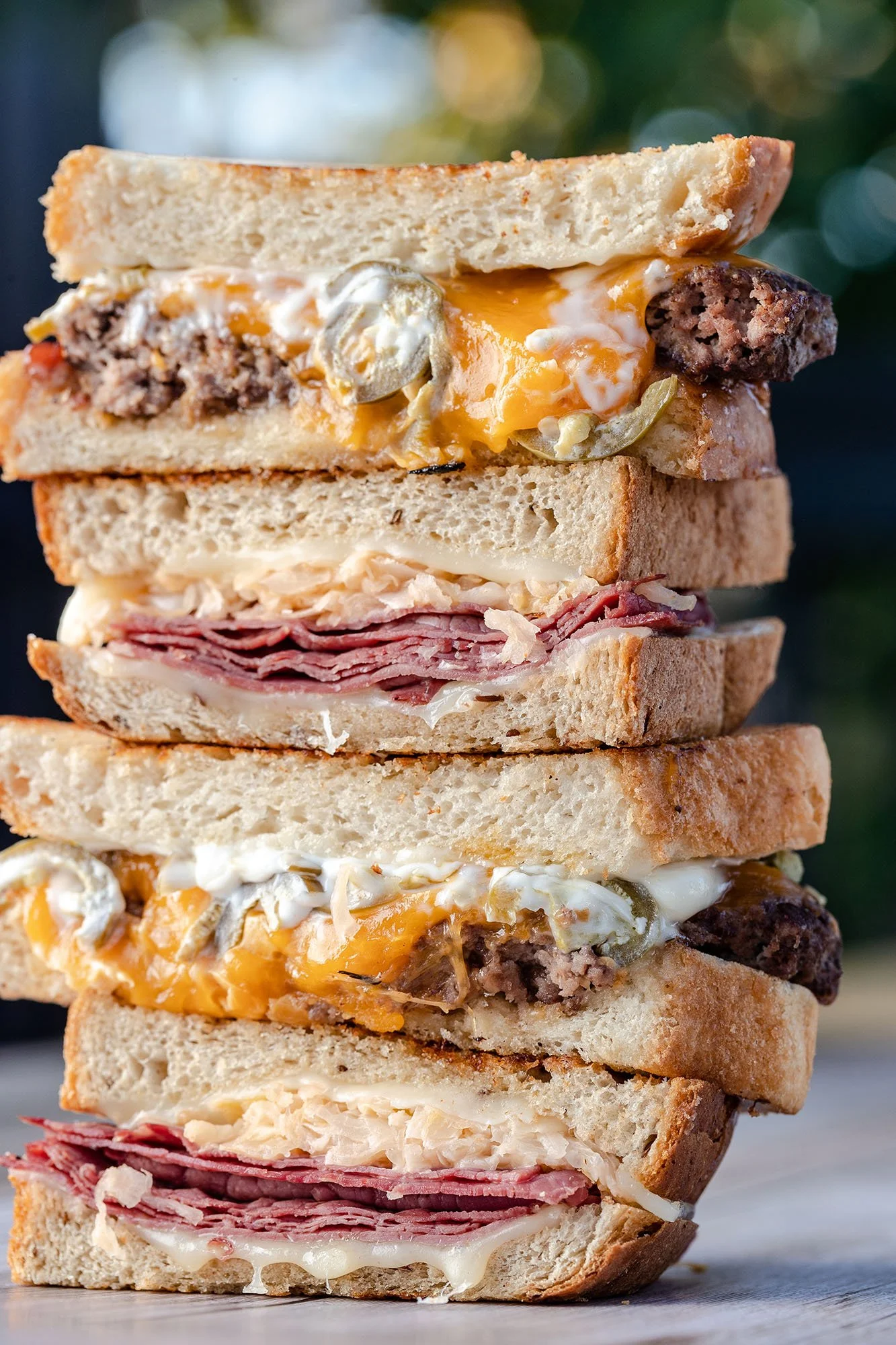 Stack of three sandwiches with different fillings including ground beef, egg, melted cheese, roast beef, and shredded chicken, with visible bread and toppings.