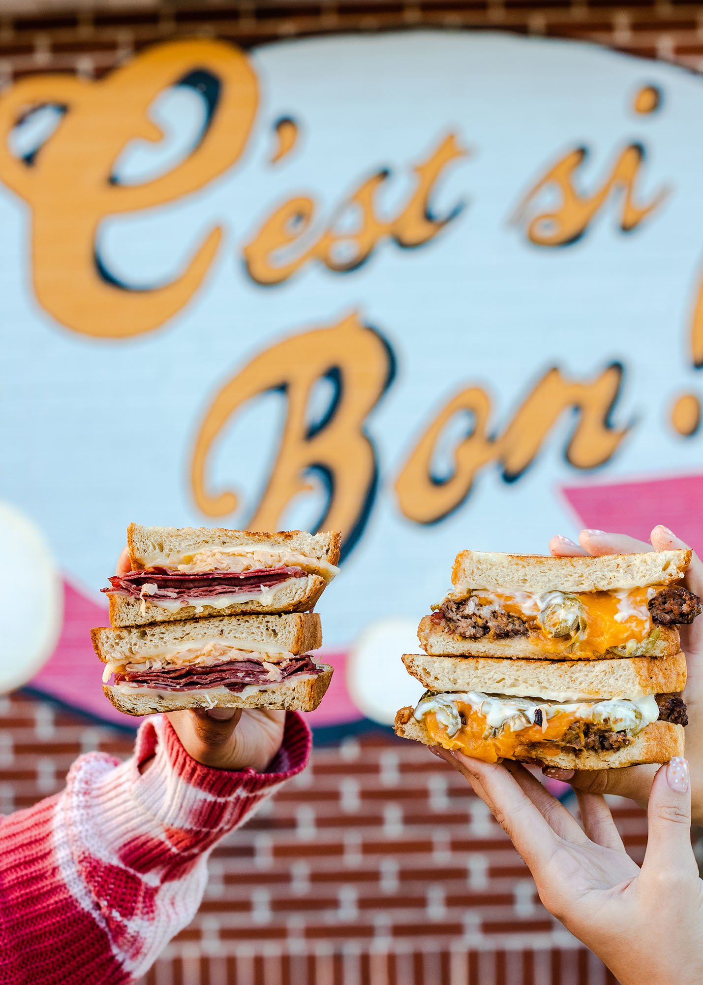 Two hands holding sandwiches in front of a sign that says 'Feed n Ban!' with a brick wall background.