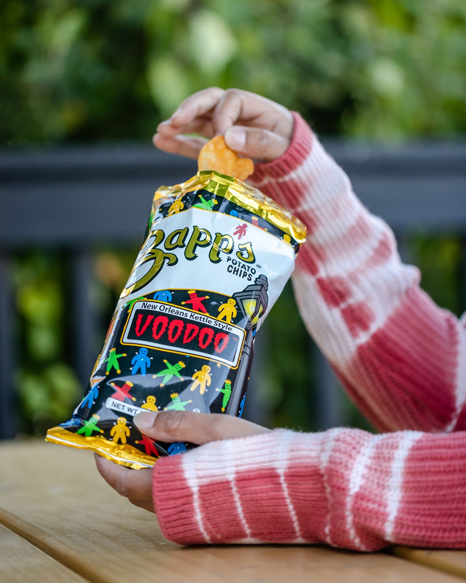 A person wearing a red and white striped sweater holding a bag of Zapp's Potato Chips labeled 'Voodoo' with a potato chip in hand.