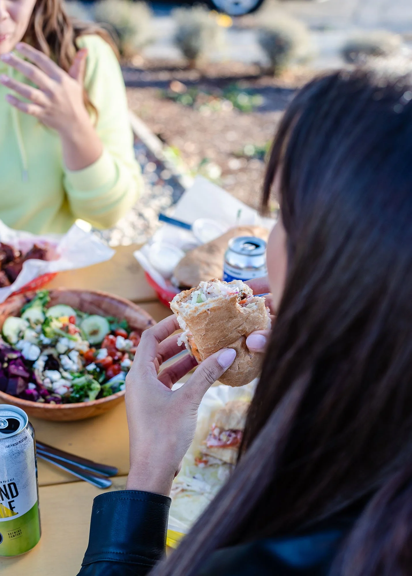 Person holding a sandwich at outdoor picnic with salad and canned beverage on table.