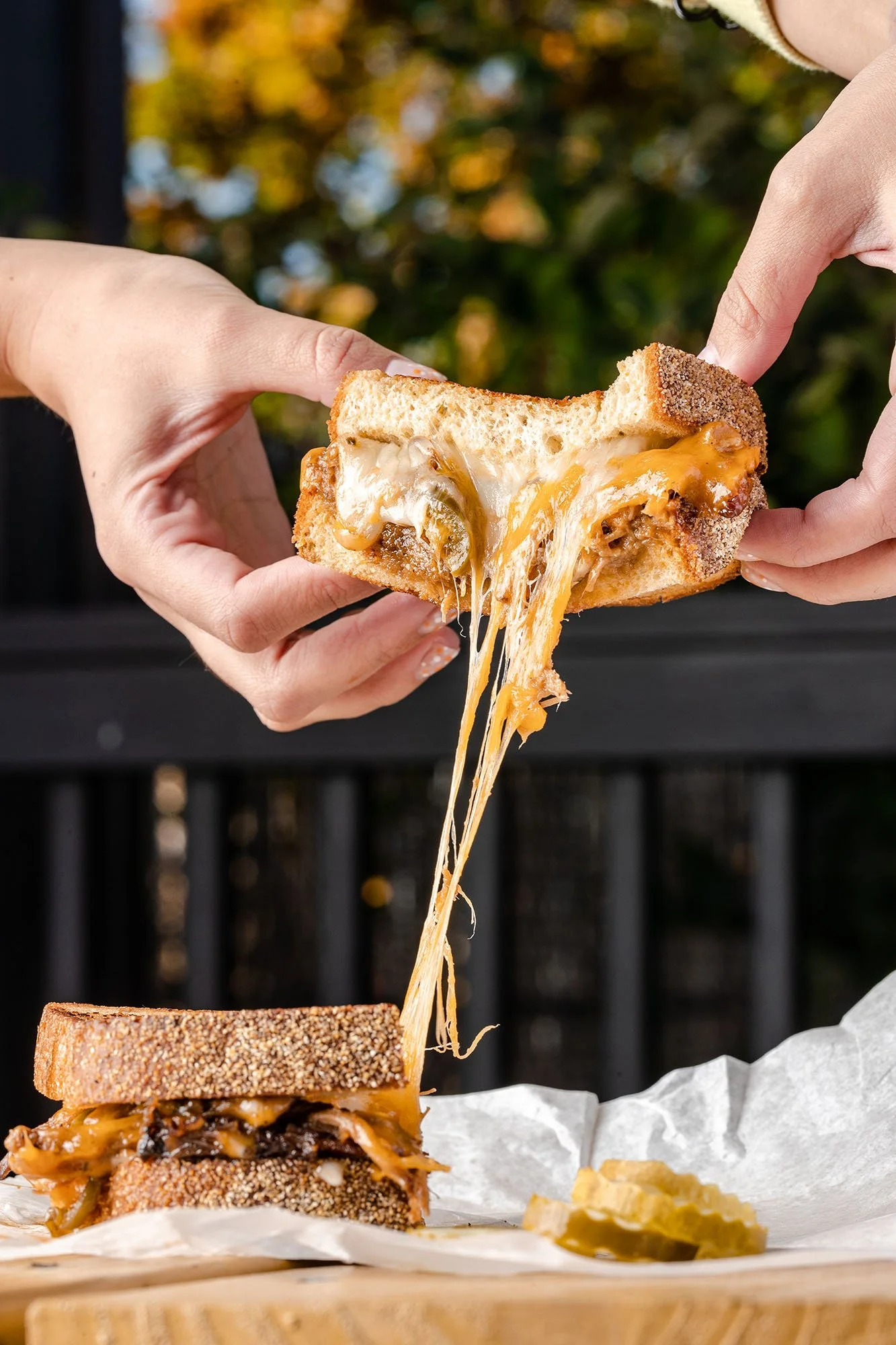 Close-up of a person pulling a cheesy sandwich apart, with melted cheese stretching between the two halves, on a wooden surface with pickles in the background.