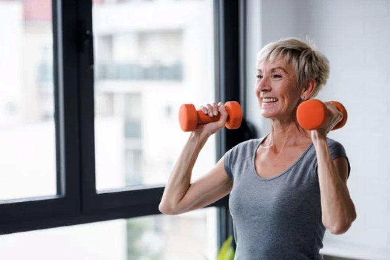 a woman over 65 wearing a day gray shirt lifts orange barbells in front of a window