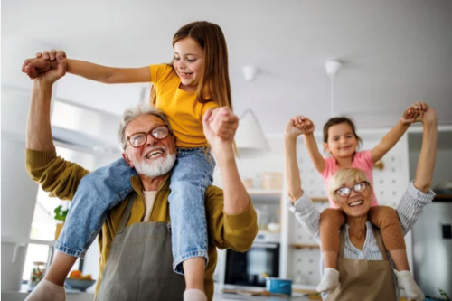 A older couple wearing aprons parade around a kitchen with their granddaughters on their shoulders. Everyone is smiling and happy