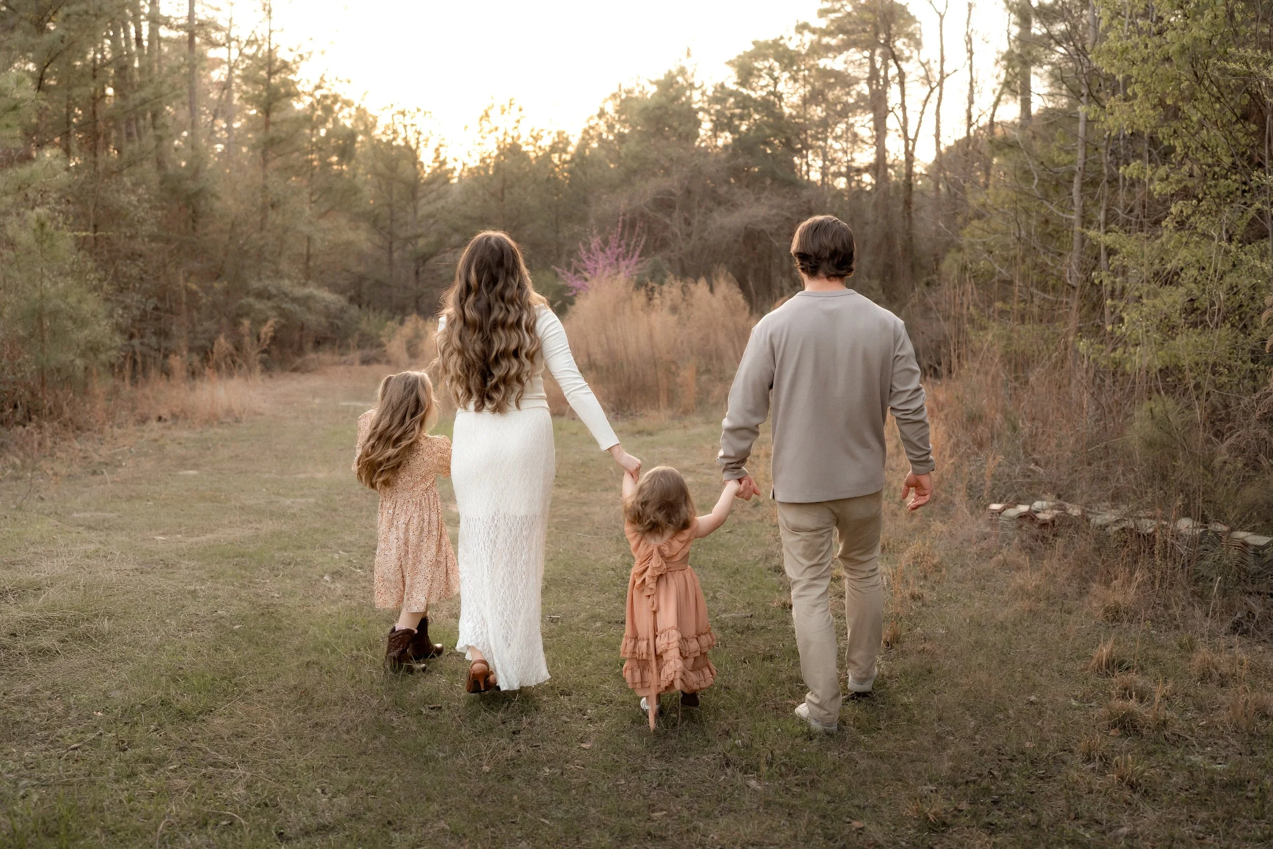 Family of four, two little girls, mother, and father, holding hands walking outside in open field surrounded by trees towards the sunset.