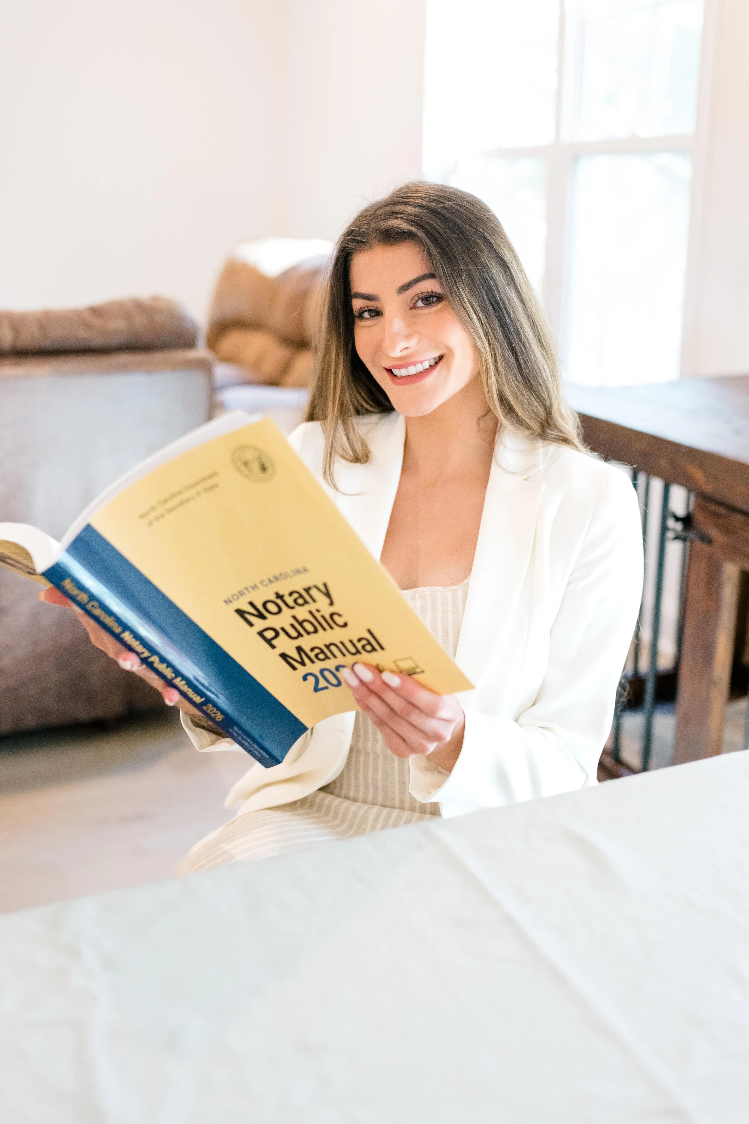 Woman wearing white jacket and tan top smiling at camera sitting at a table while holding open yellow and blue notary public manual in both hands