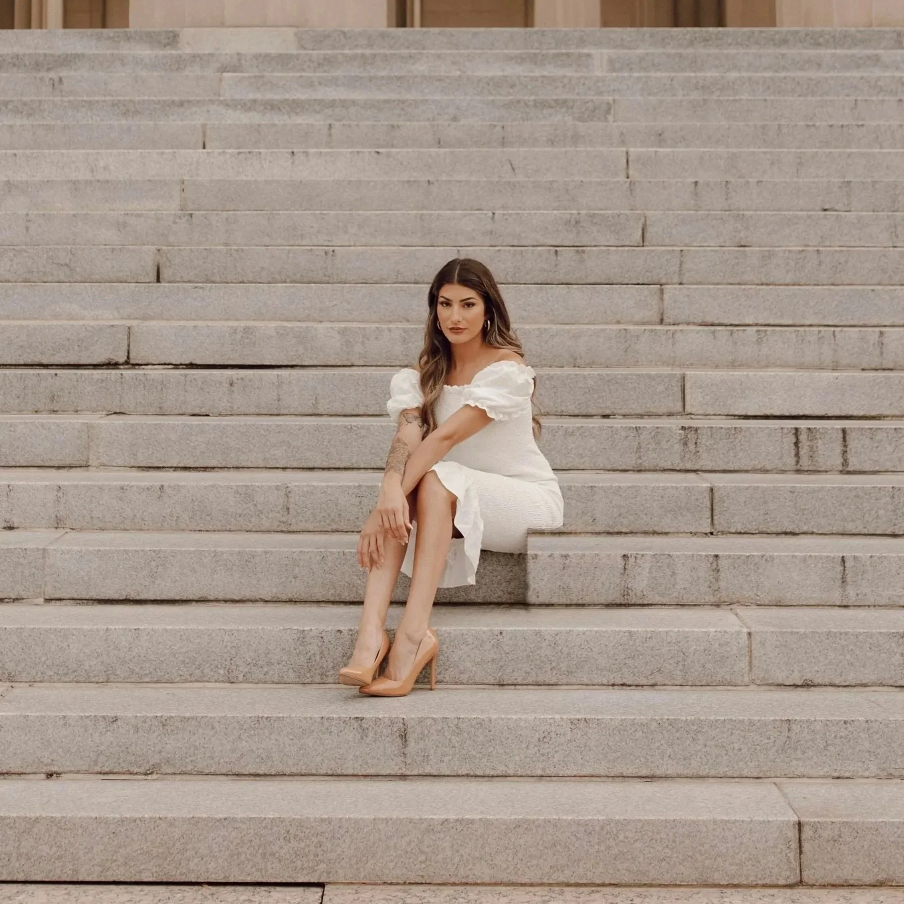 Woman in white dress sitting on row of steps with hands crossed over extended legs.