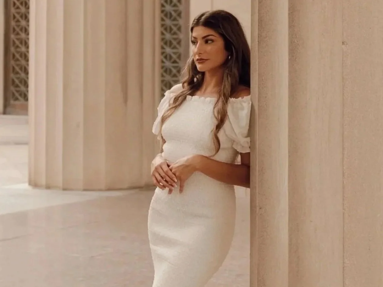 Woman wearing white dress, with hands held across waist, leaning against white pillar