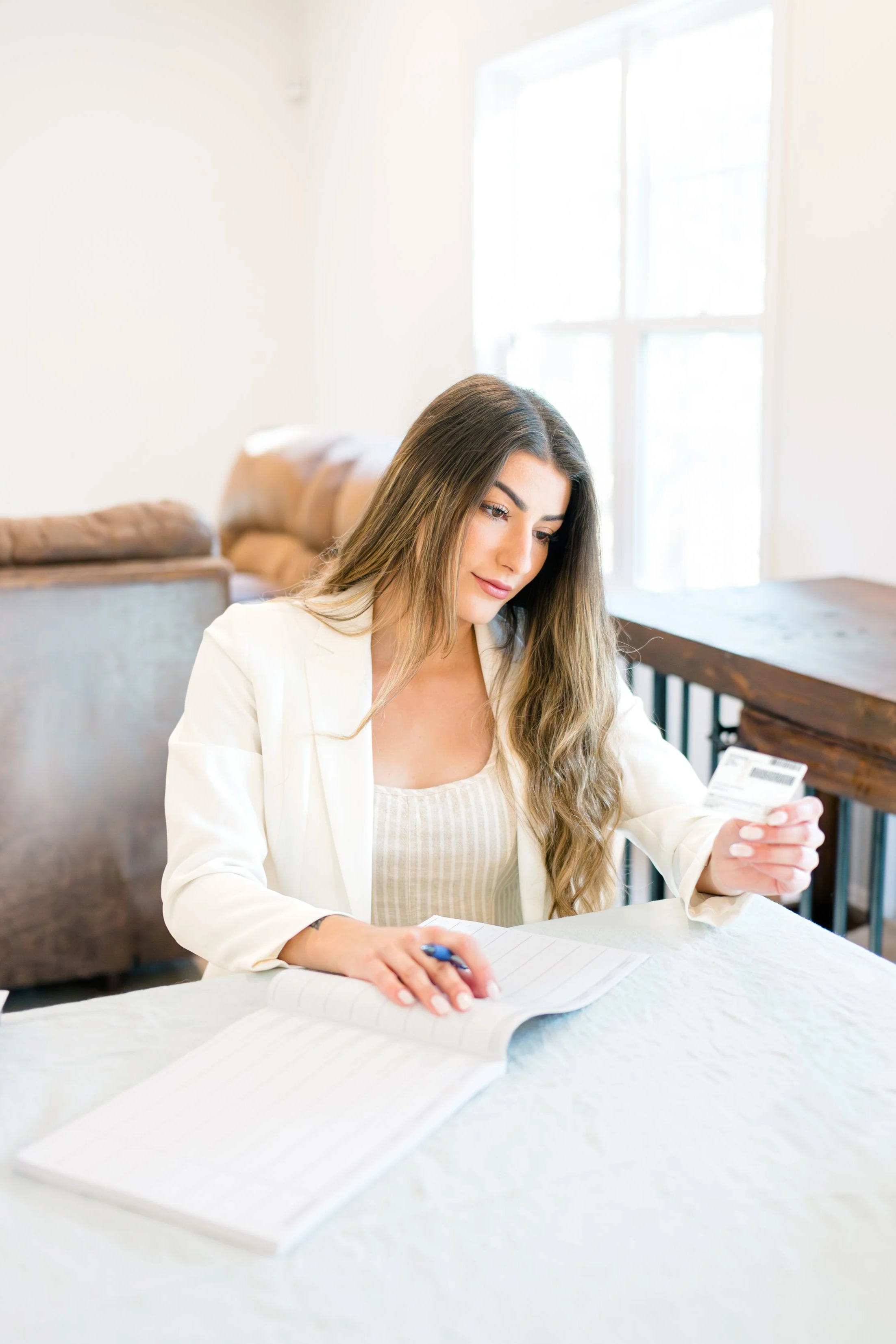 Woman wearing white jacket and tan top looking at a driver's license held in left hand and right hand holding pen and resting on table where woman is sitting