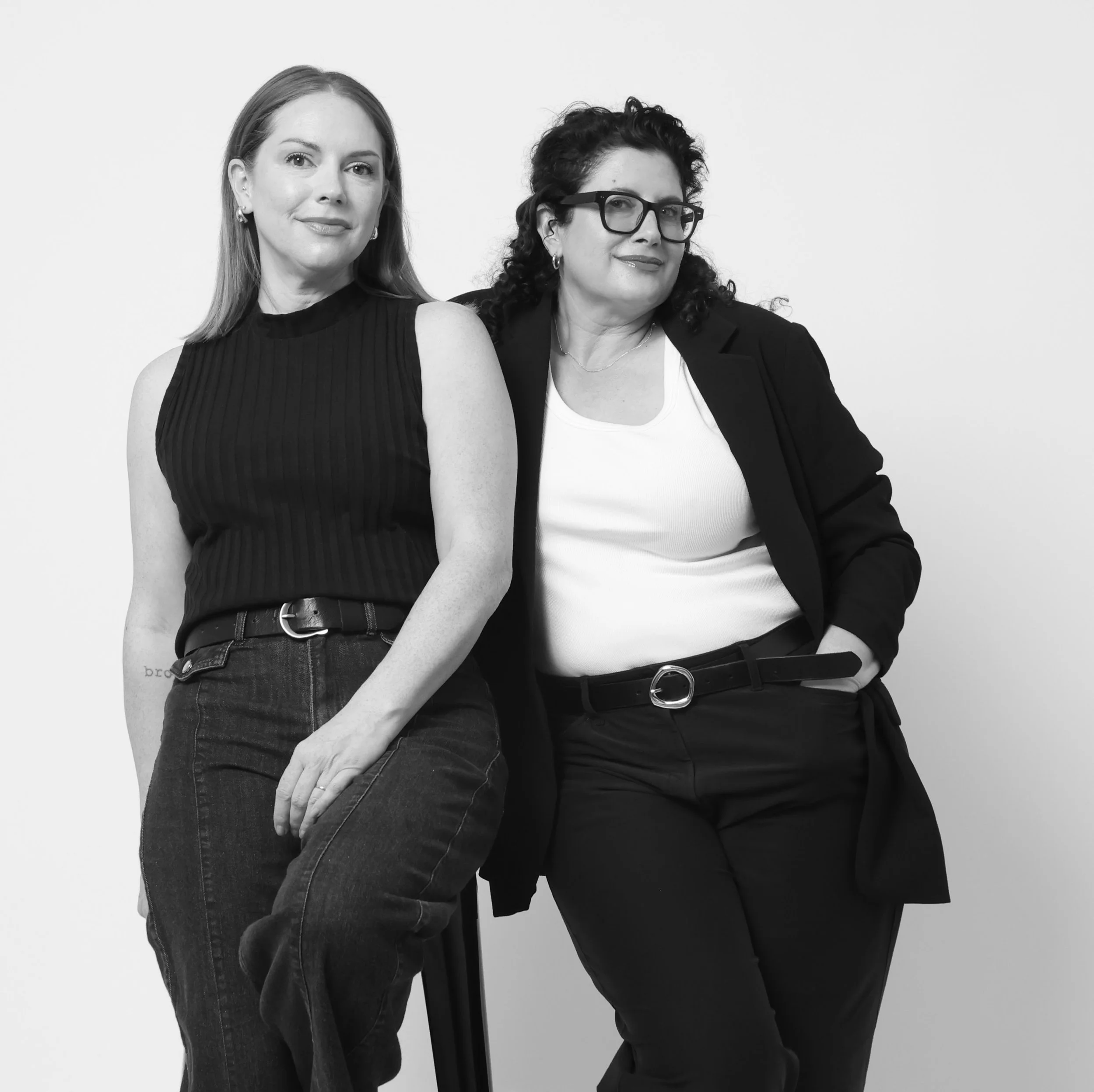 Two women standing close together, one with long hair wearing a sleeveless top and jeans, the other with curly hair, wearing glasses and a blazer, against a plain background.