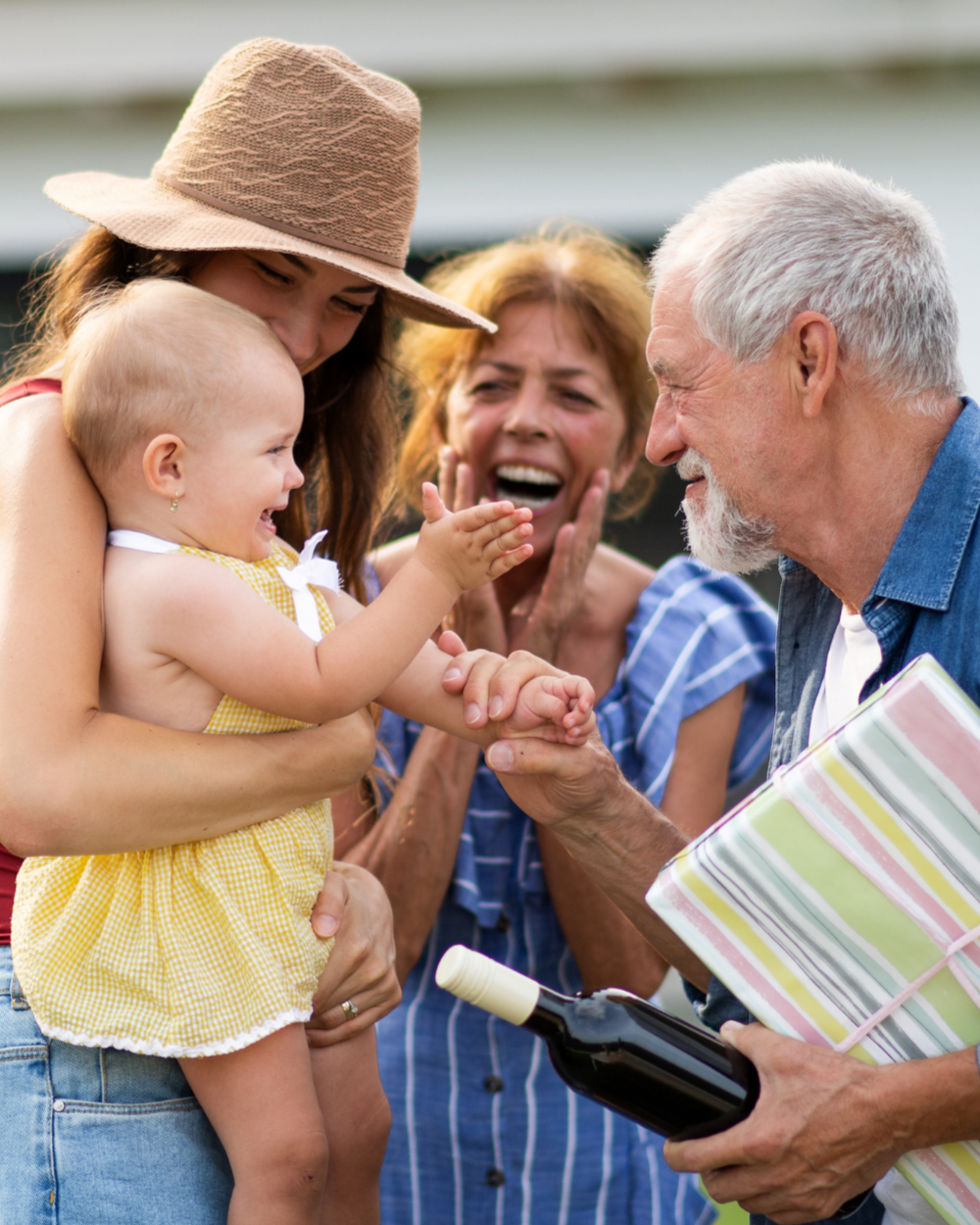 Family celebrating a birthday outdoors, with an elderly man giving a gift and a bottle of wine to a smiling baby girl, family members clapping and smiling.