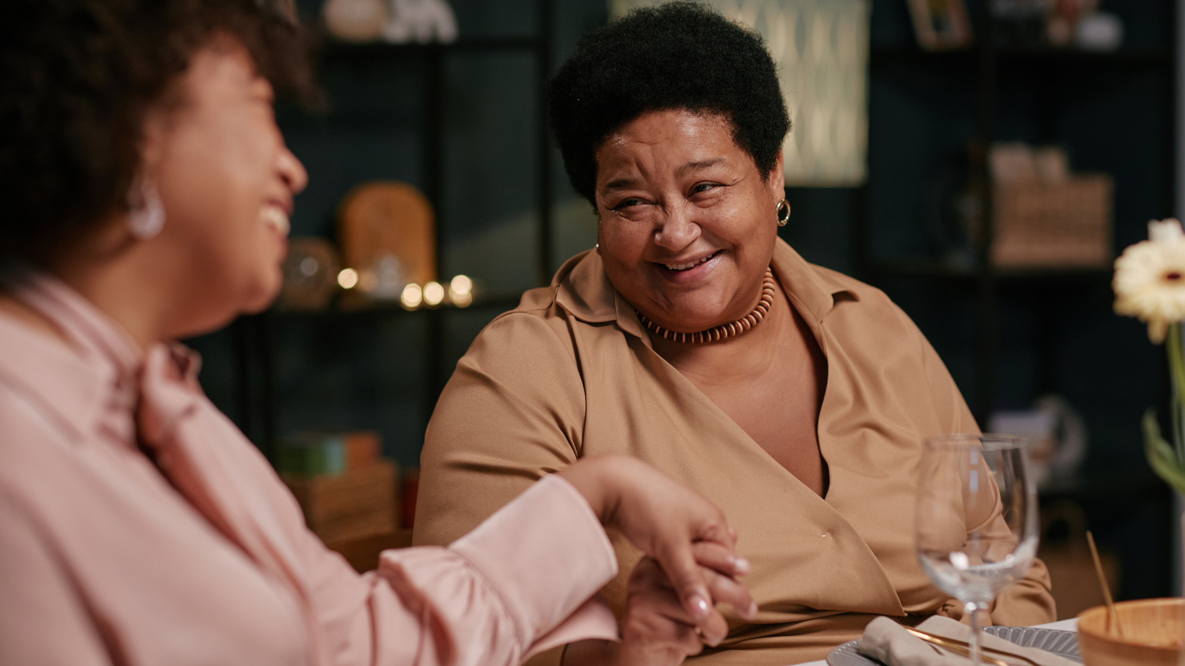 Two women are sitting at a table, holding hands, and smiling at each other in a warmly lit room with dark colored shelves in the background.