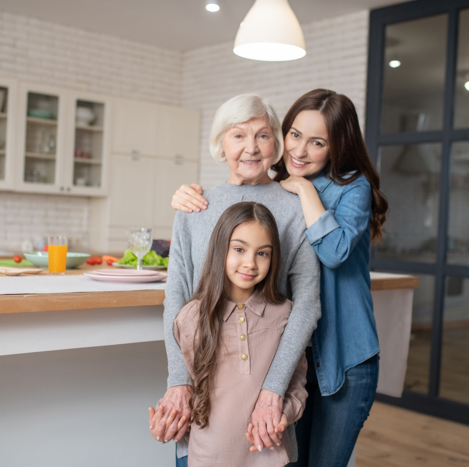 Three generations of women hugging in a modern kitchen with a breakfast bar, plates, glasses, and a salad. A grandmother, mother, and young girl smiling together.