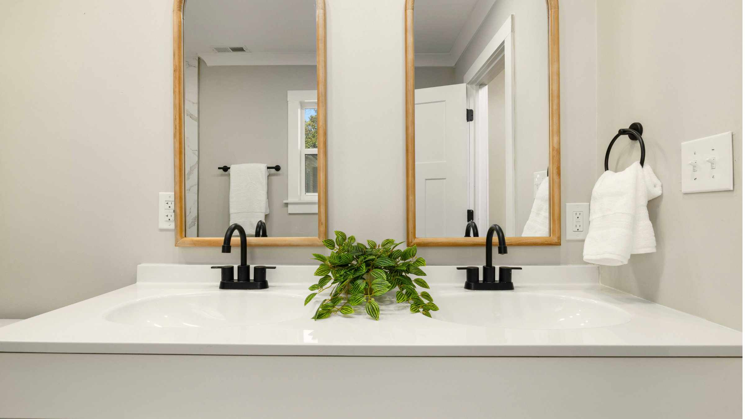Bathroom vanity with two black faucets, a plant, and three large mirrors with wooden frames, white walls, a towel on a black towel bar, and a window reflected in the mirror.