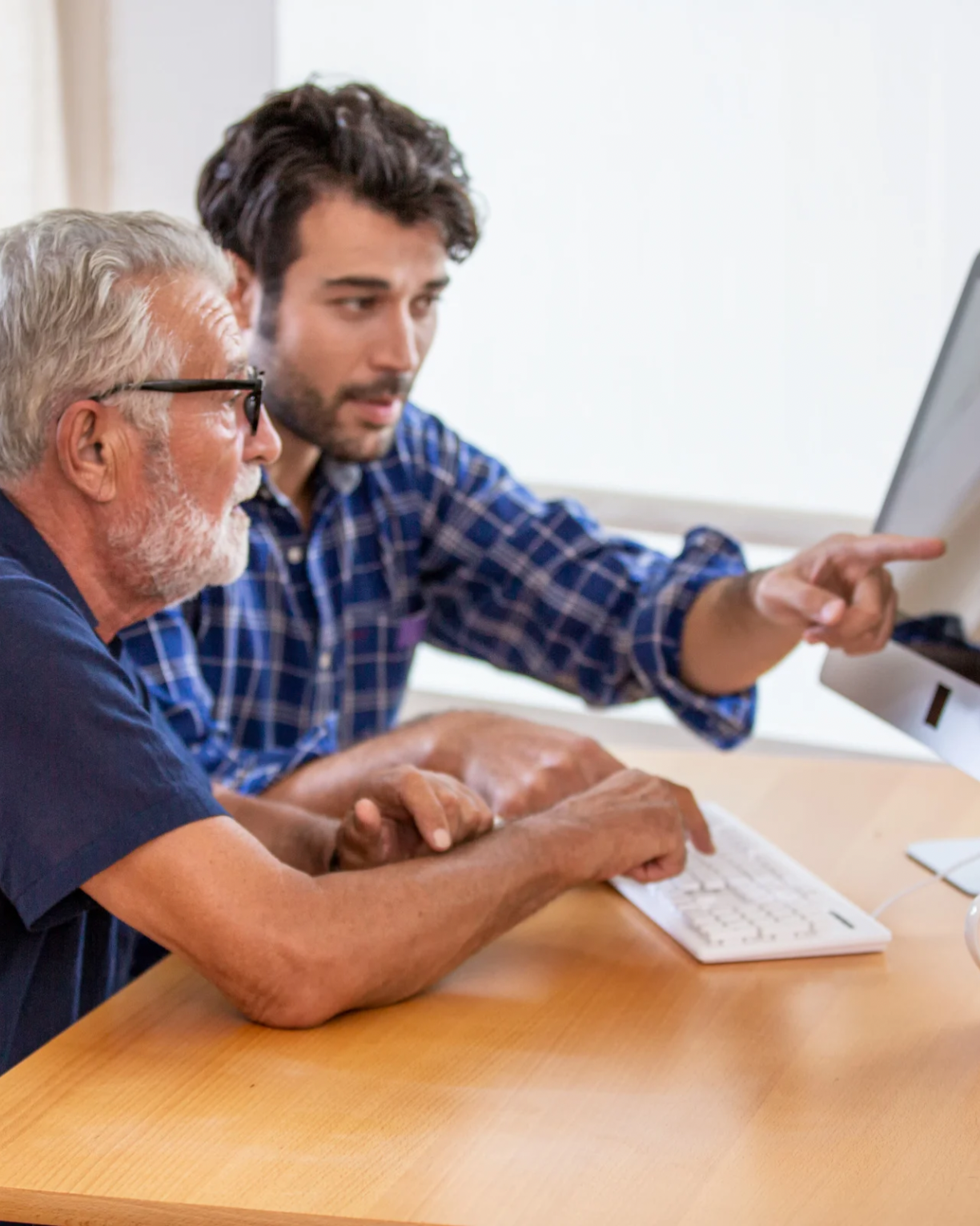 Two men, one older with gray hair and glasses and one younger, sitting at a desk working together on a computer. The younger man is pointing at the screen.