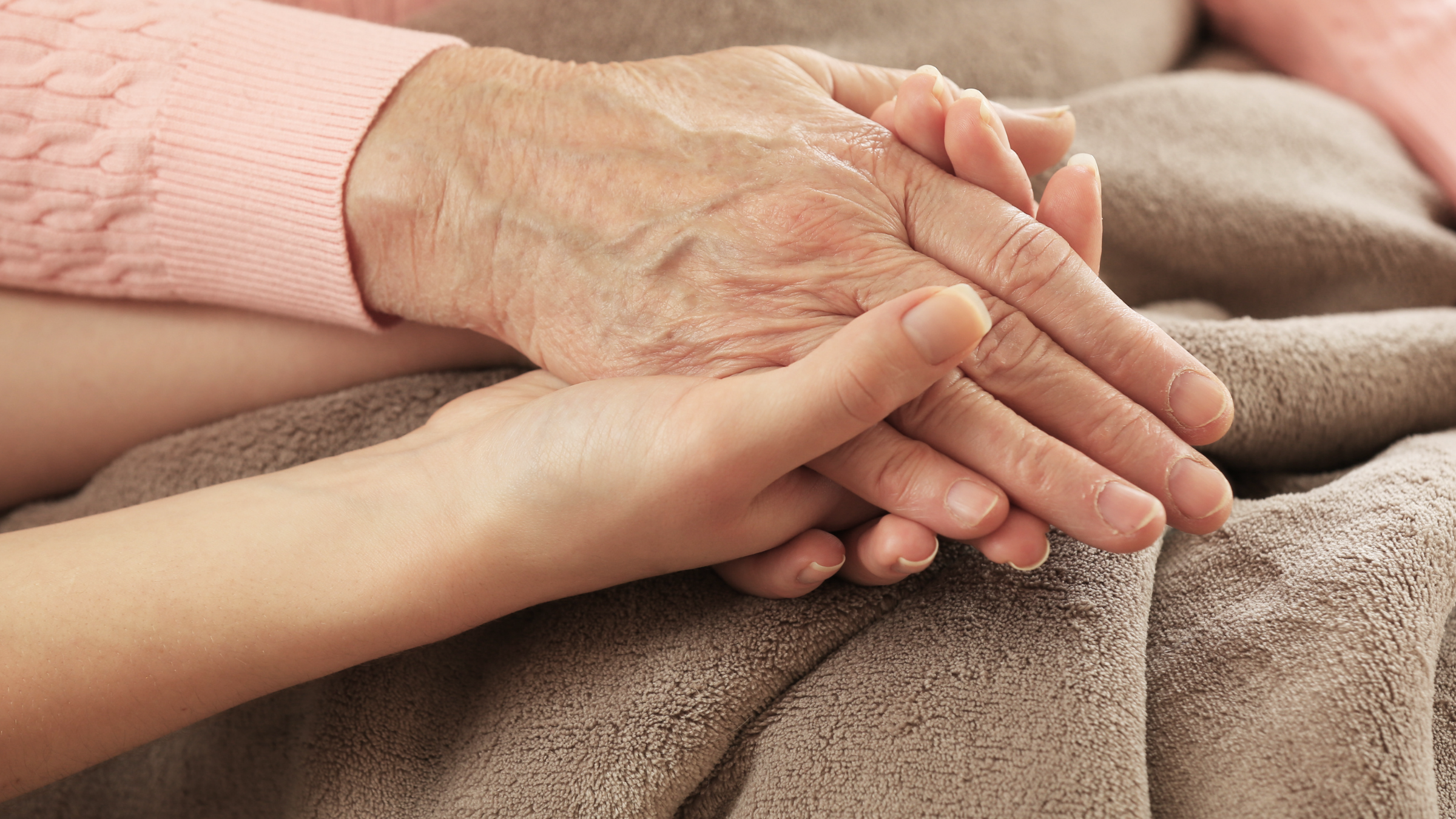 Two hands, one older with wrinkles, holding a younger, smooth hand, both resting on a soft brown fabric surface.