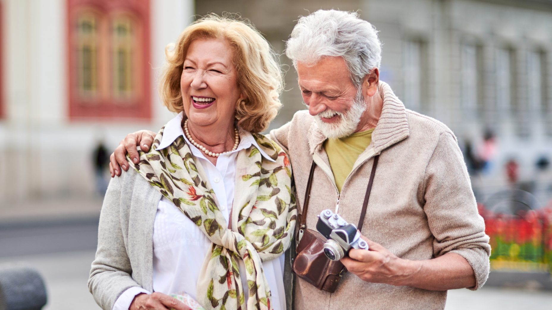 An elderly man and woman smiling and sharing a happy moment, with the man showing a camera to the woman, outdoors in a city setting.