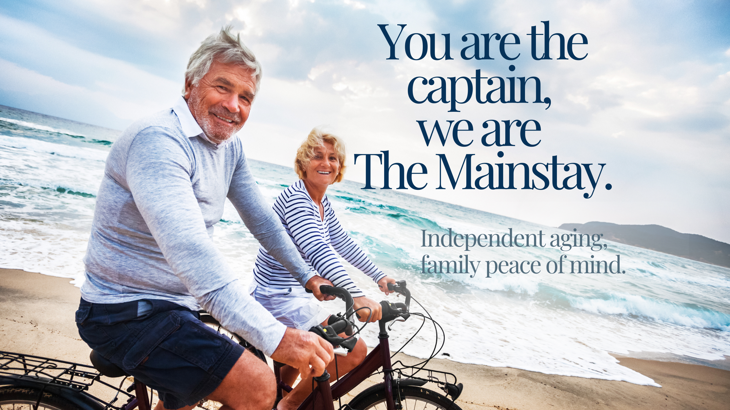 Older man and woman smiling while riding bikes on the beach near the ocean, with a cloudy sky in the background.
