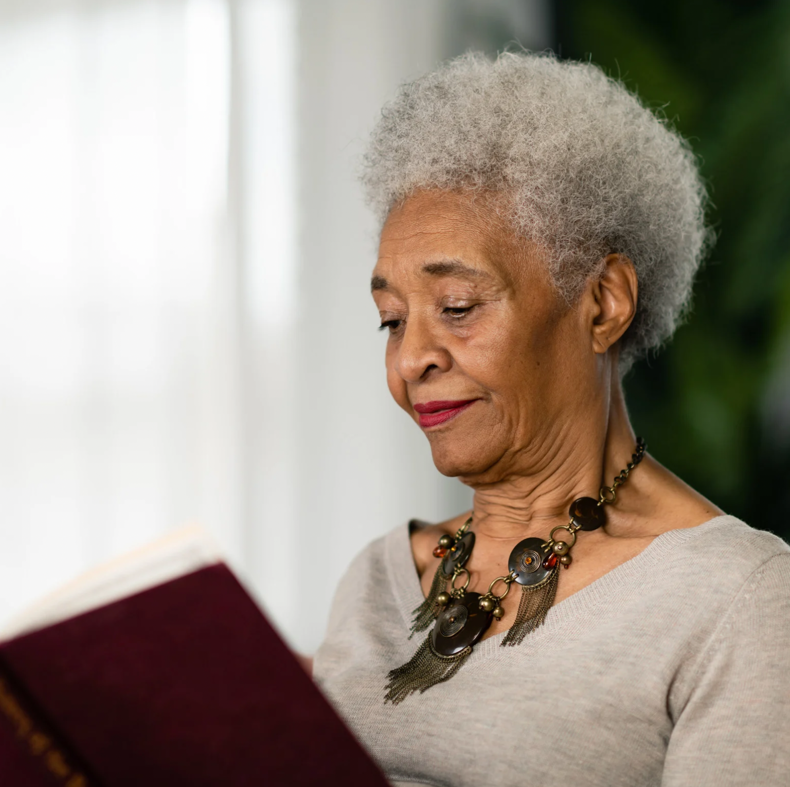 An elderly woman with white, curly hair reading a book, wearing a beige top and a statement necklace with large beads and tassels.