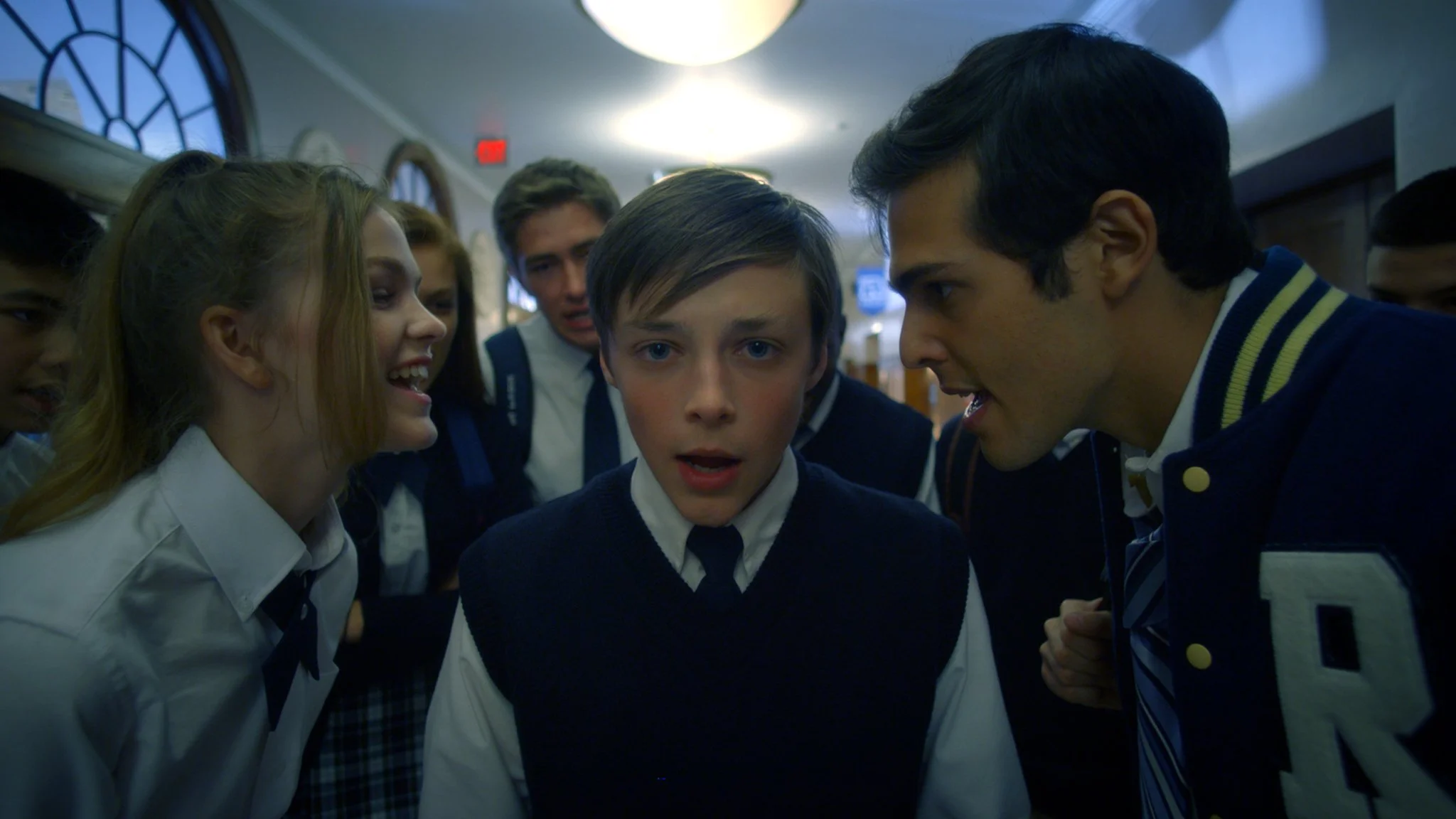 A group of students in school uniforms crowded around a boy in a hallway, looking at him with concerned and curious expressions.