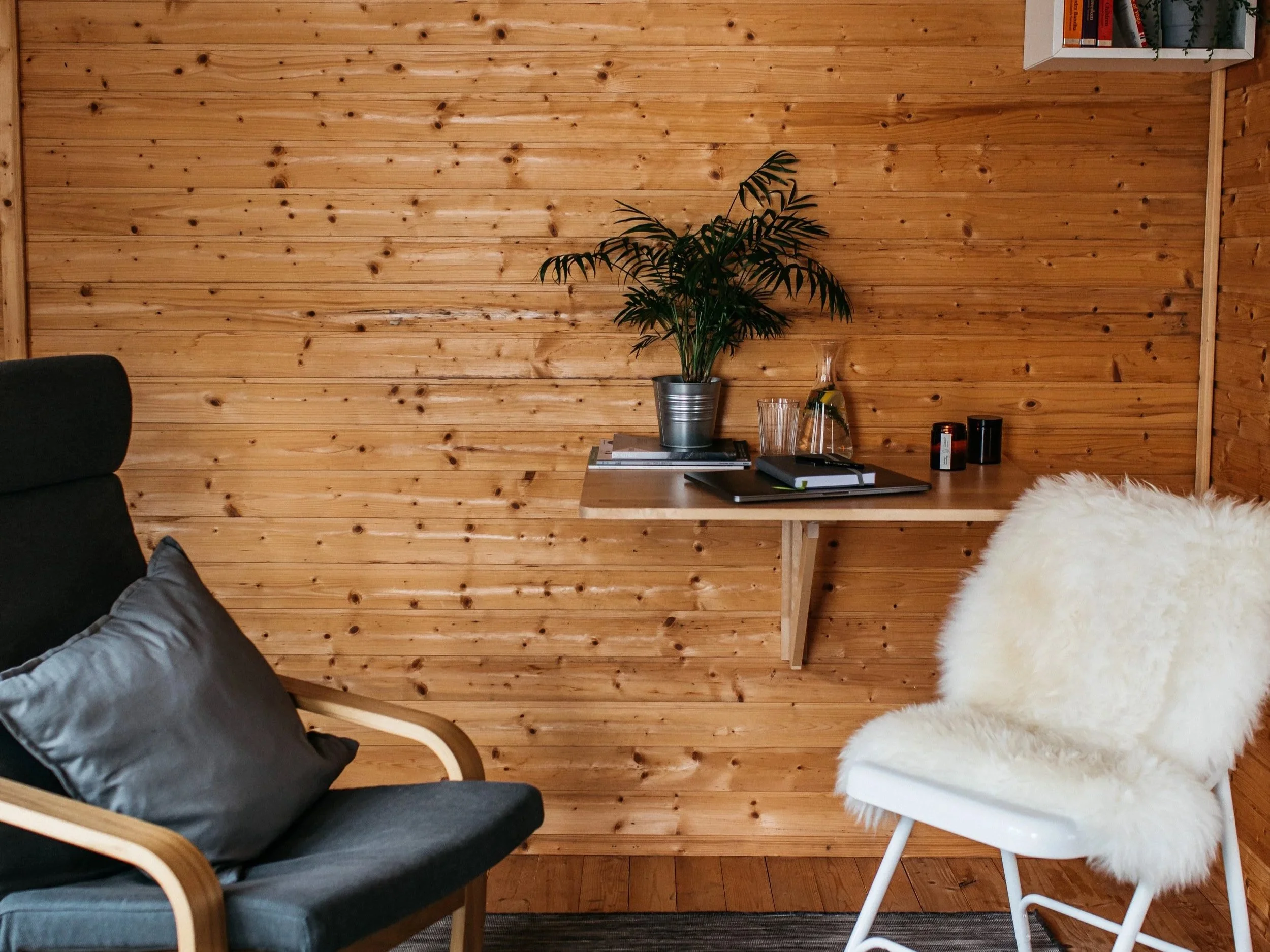 A cozy room with a wooden wall, a black armchair with a gray pillow, a white fur-covered chair, and a small floating desk with a potted plant, glassware, notebooks, and some small containers.