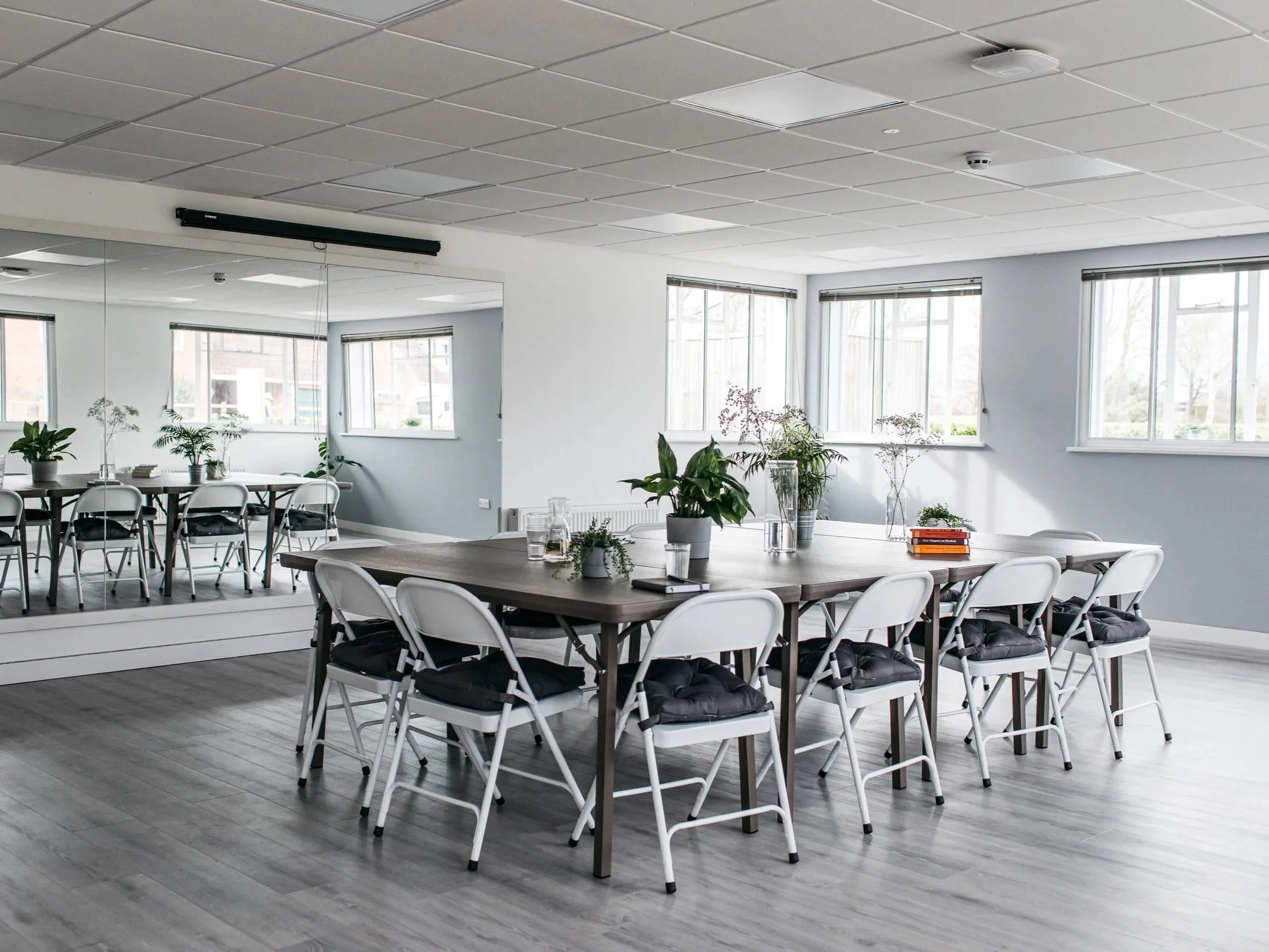 A bright meeting room with a large wooden table surrounded by white chairs with black cushions. There are plants in the center and on the windowsills, with large windows letting in natural light.