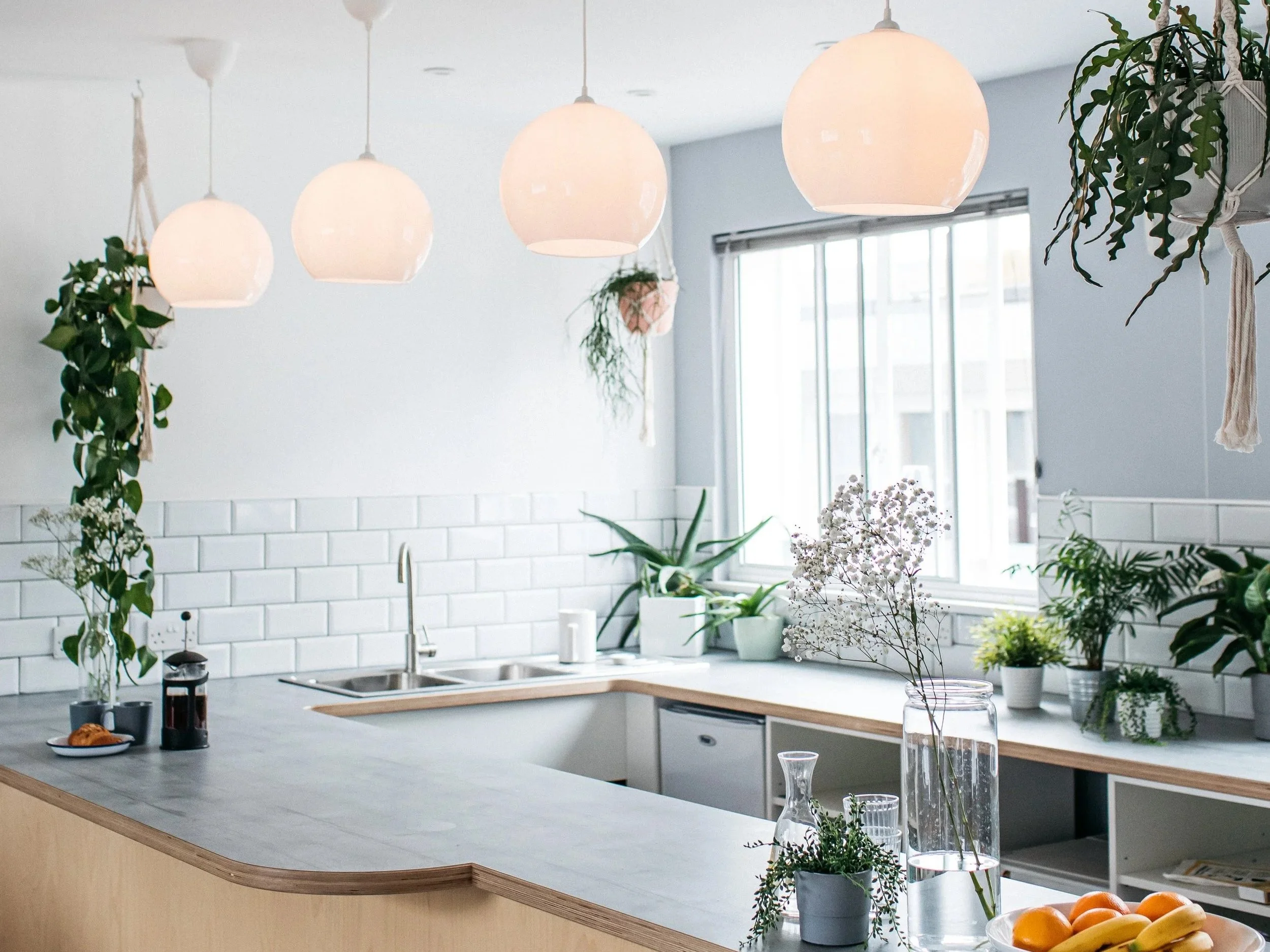 Bright kitchen with hanging pendant lights, white subway tile backsplash, white walls, and various potted plants on the countertop and near the window.