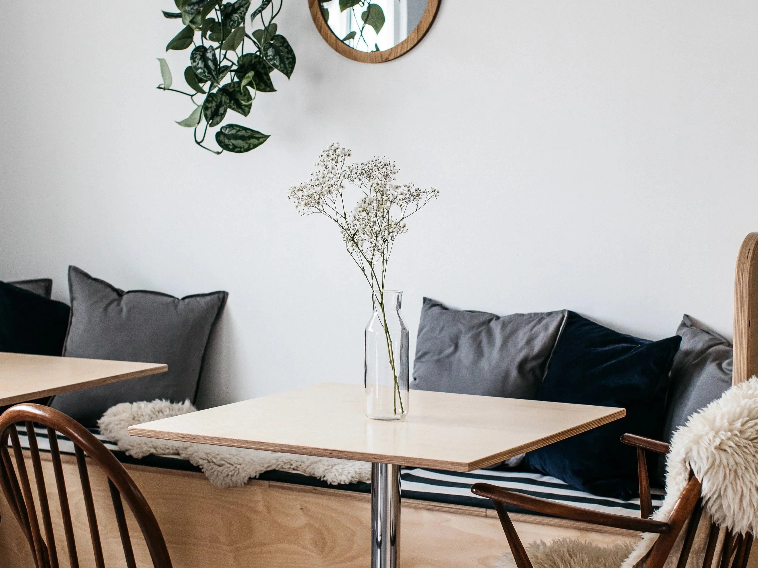 A cozy dining area with a light wood table, a clear vase with white baby's breath flowers, dark and grey cushions on a bench, a wooden chair with a throw, and a wall mirror with a round wooden frame.