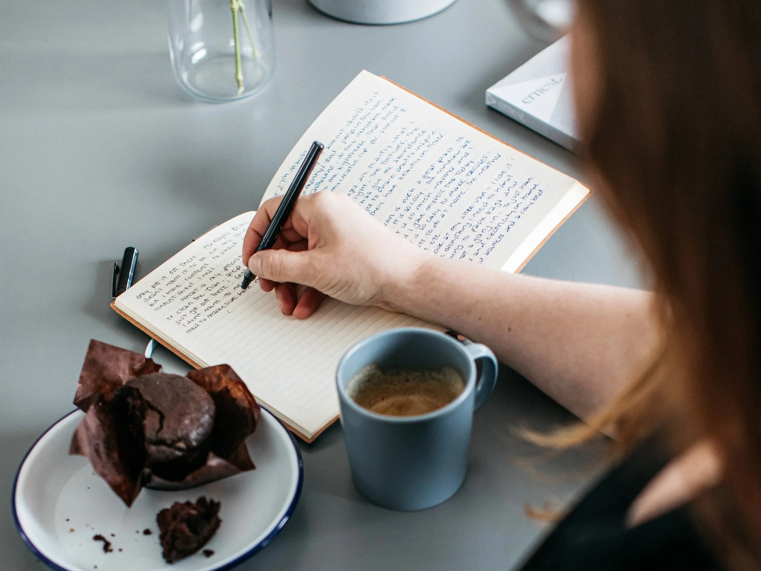 Person writing in a notebook at a table with a mug of coffee, and a plate with chocolate muffins.