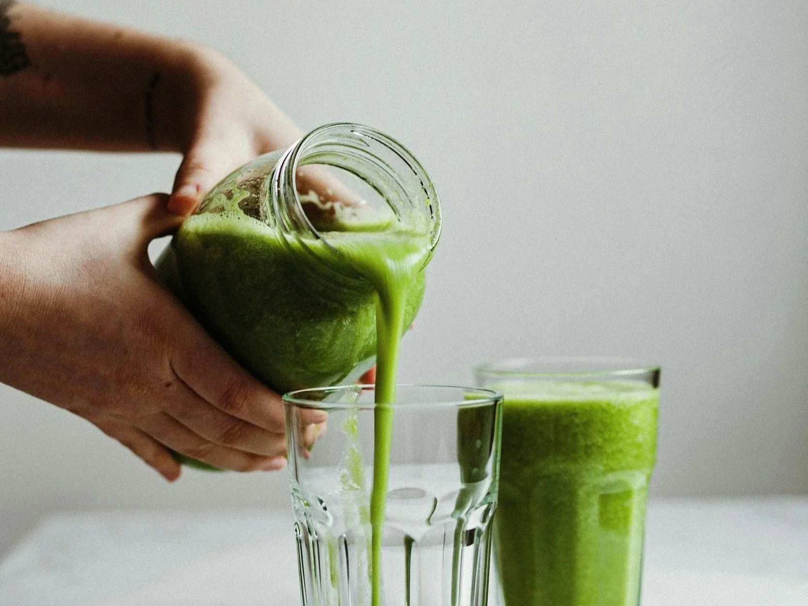 Person pouring green smoothie from jar into a glass, with a second glass of green smoothie on a white surface.