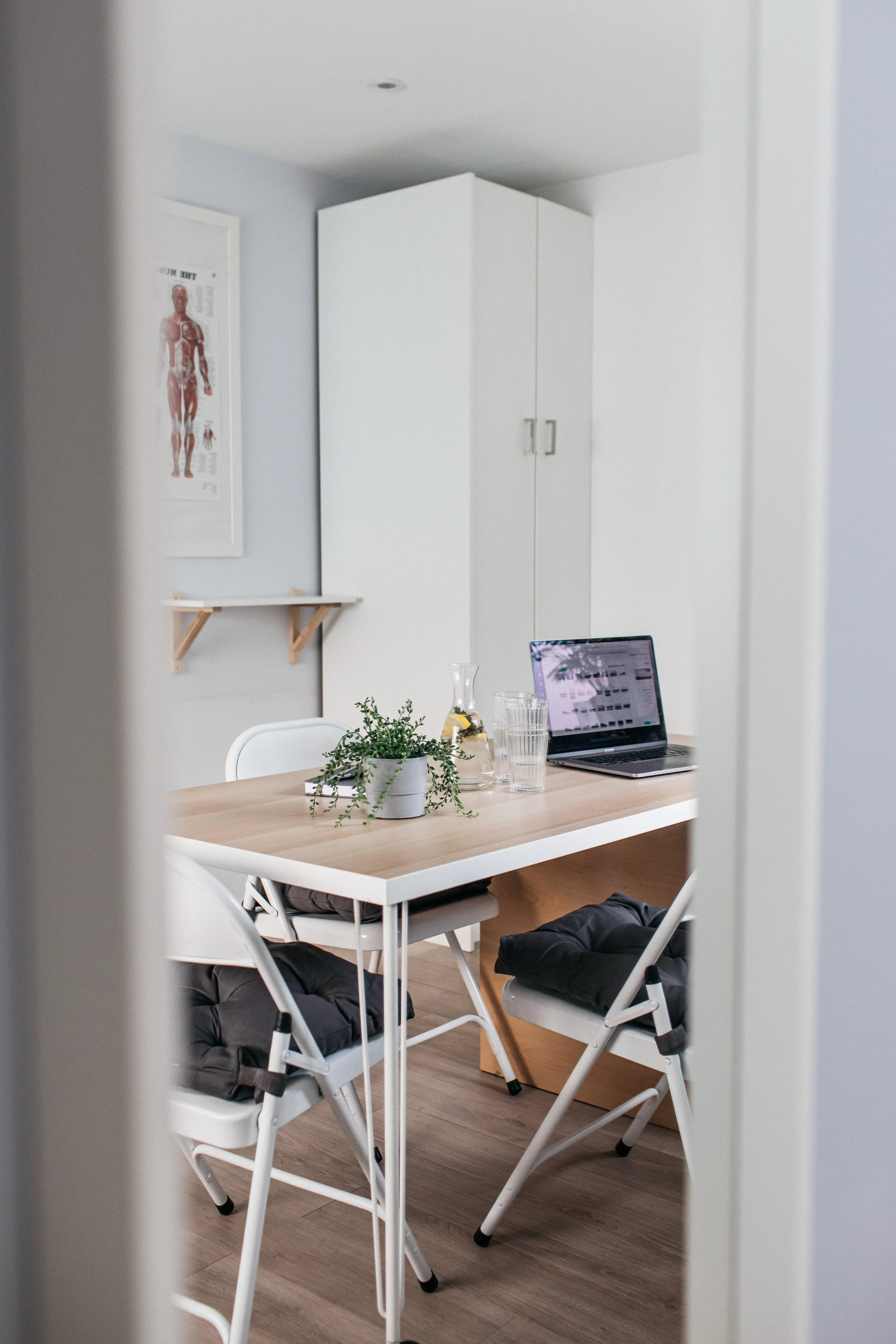 A room with a wooden table, two folding chairs, a potted plant, a water pitcher, three glasses, and an open laptop, viewed through a doorway.