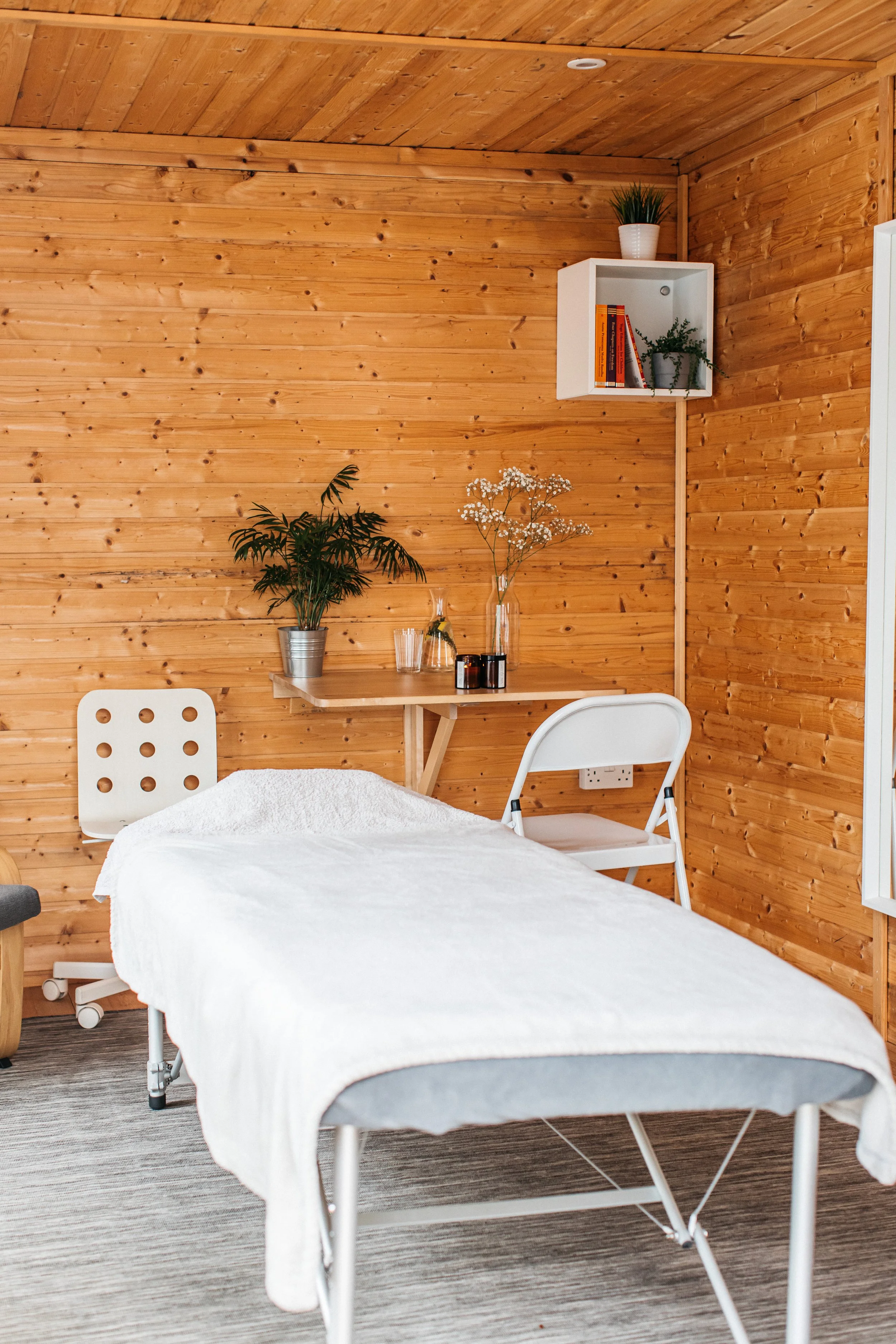 A room with a massage table covered with a white towel, a white chair, a small wooden desk with plants and flowers, a white wall-mounted shelf with books and a small plant, and wooden walls and ceiling.