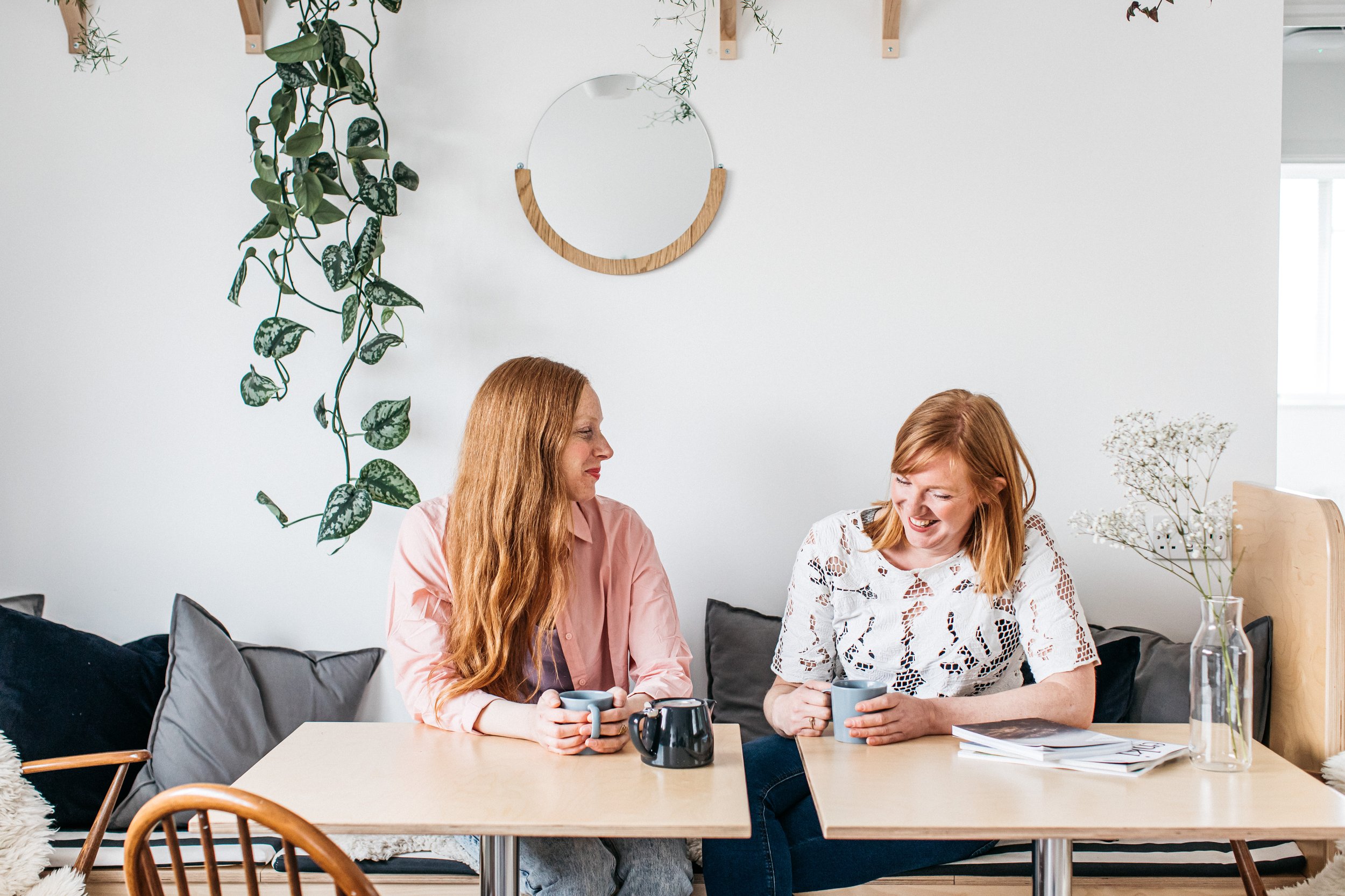Two women talking and smiling while sitting at a table with cups, a pitcher, and some papers in a cozy, decorated room with plants and a mirror.