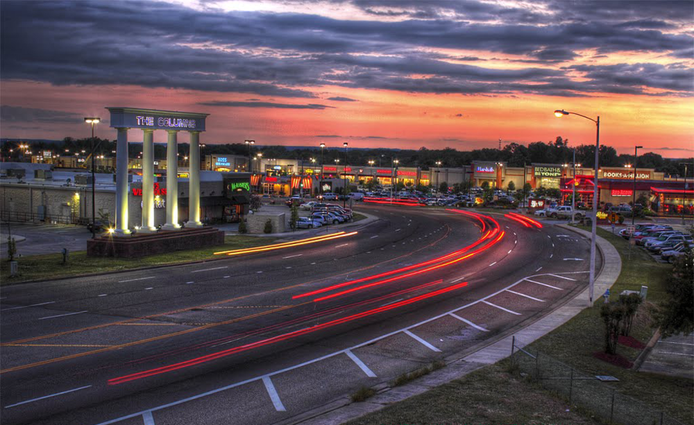 A shopping center parking lot during sunset with light trails from moving vehicles, a large illuminated sign that reads 'The Colony', and stores visible in the background.