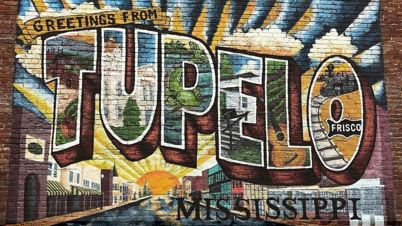 Street art mural with the words 'Greetings from Tupelo Mississippi' in large colorful letters, featuring images of houses, trees, a train, and a sunrise.