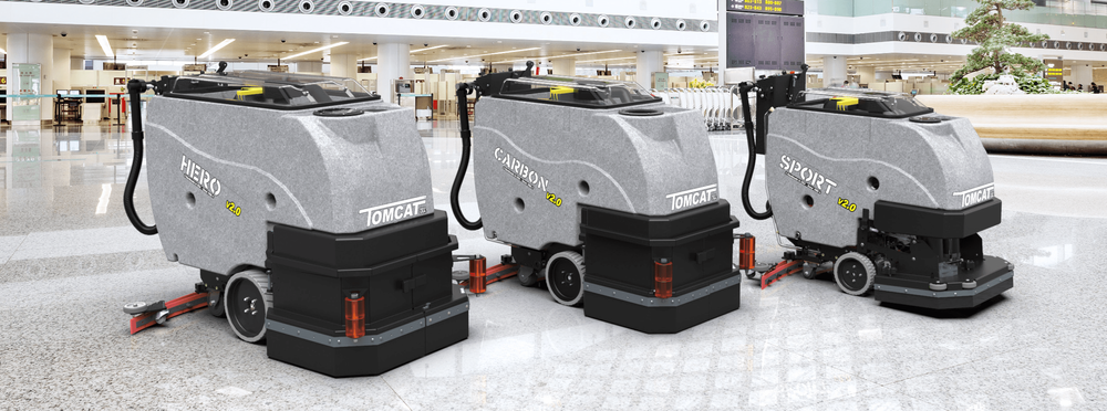 Three large autonomous floor cleaning robots with gray bodies, labeled 'HERO', 'CARRIER', and 'SPORT', are cleaning an airport terminal floor.