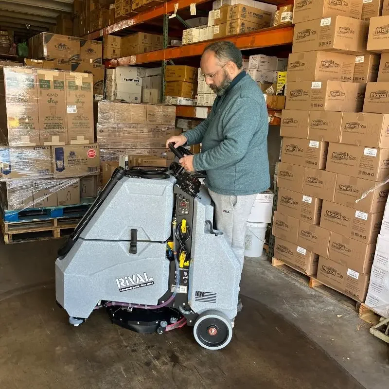 A man operating a commercial floor cleaning machine in a warehouse surrounded by stacked cardboard boxes.