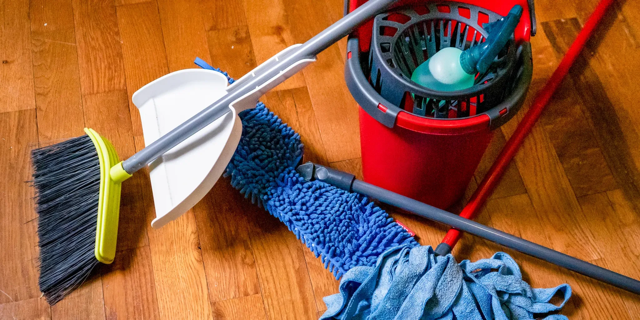 Cleaning supplies on a hardwood floor, including a dustpan and broom, a mop with a blue microfiber pad, a bucket with a spray bottle inside, and a cloth.