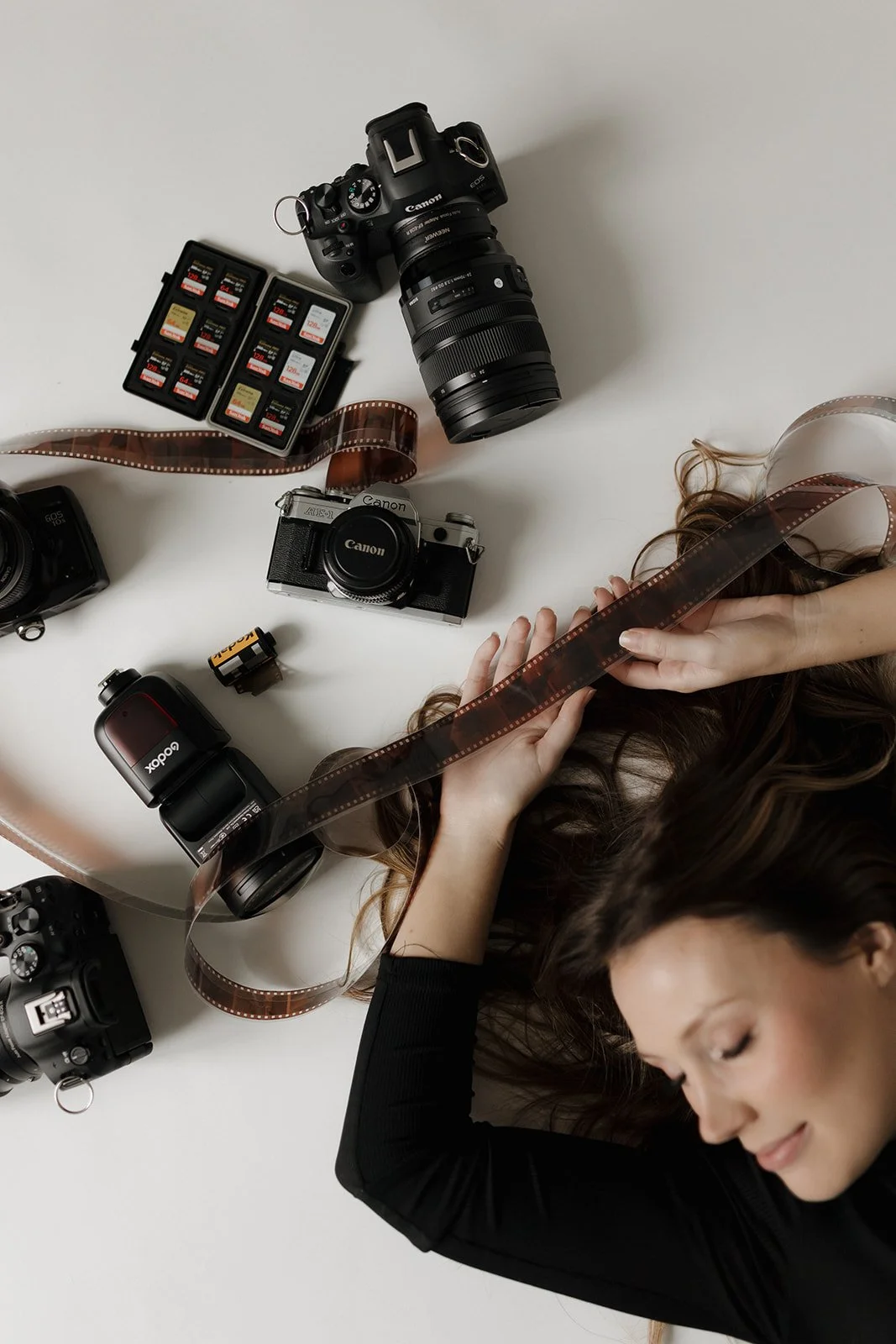 A woman with long brown hair is lying down on a white surface, holding a strip of film in her hands surrounded by photography equipment including cameras, lenses, memory cards, batteries, and a person’s hand in the corner.