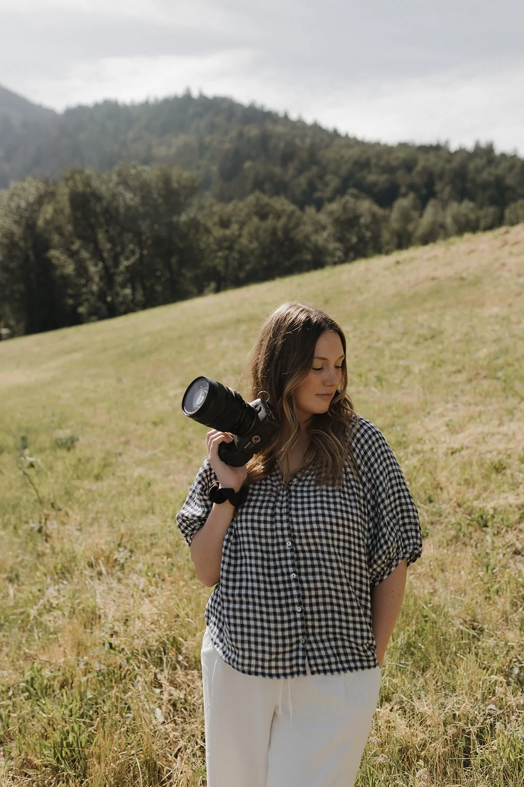 A young woman standing in a grassy field holding a camera on her shoulder, with forested hills in the background.