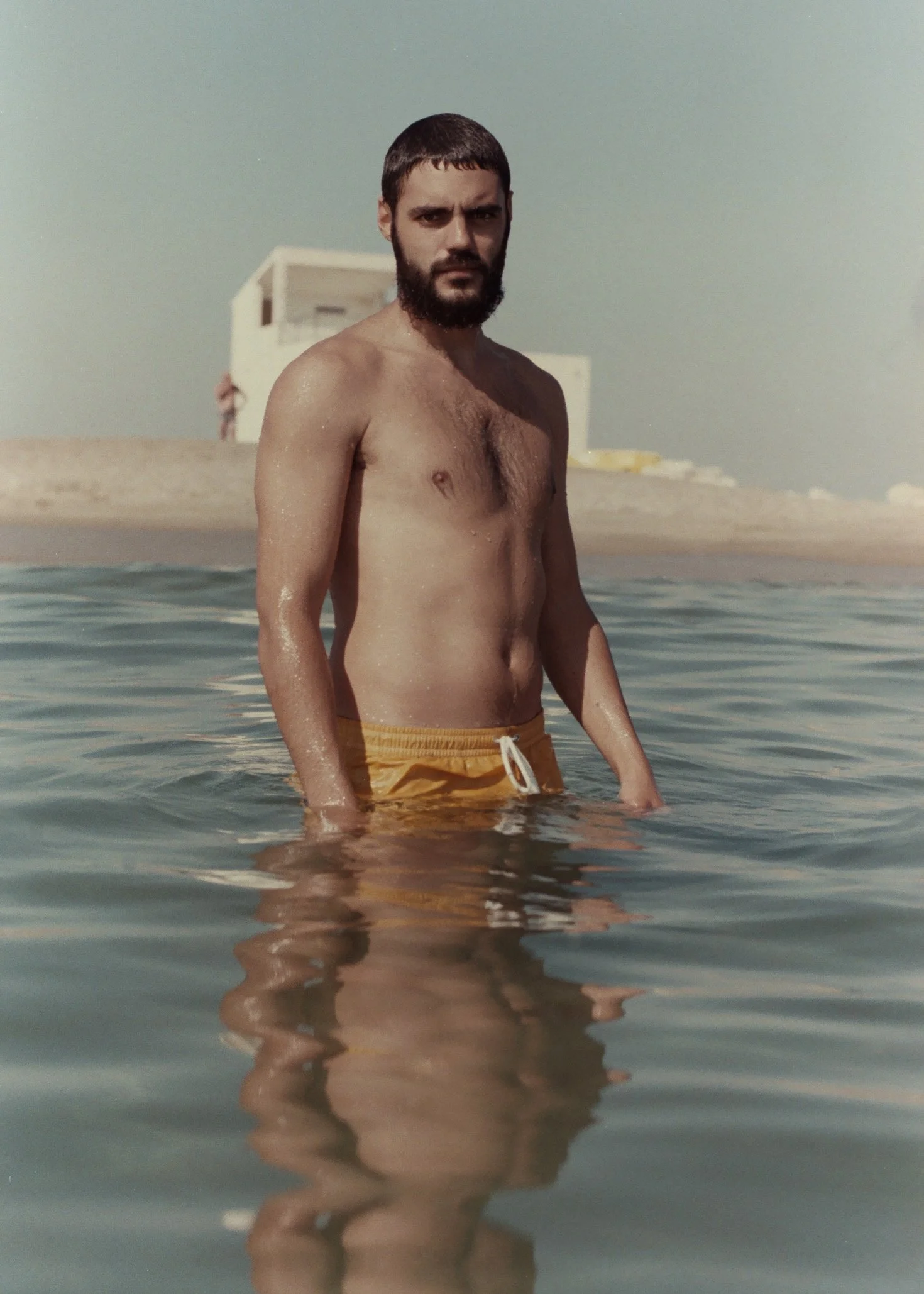 Un homme nu portant un short jaune, debout dans l'eau de mer, avec des bâtiments en arrière-plan sur la plage.
