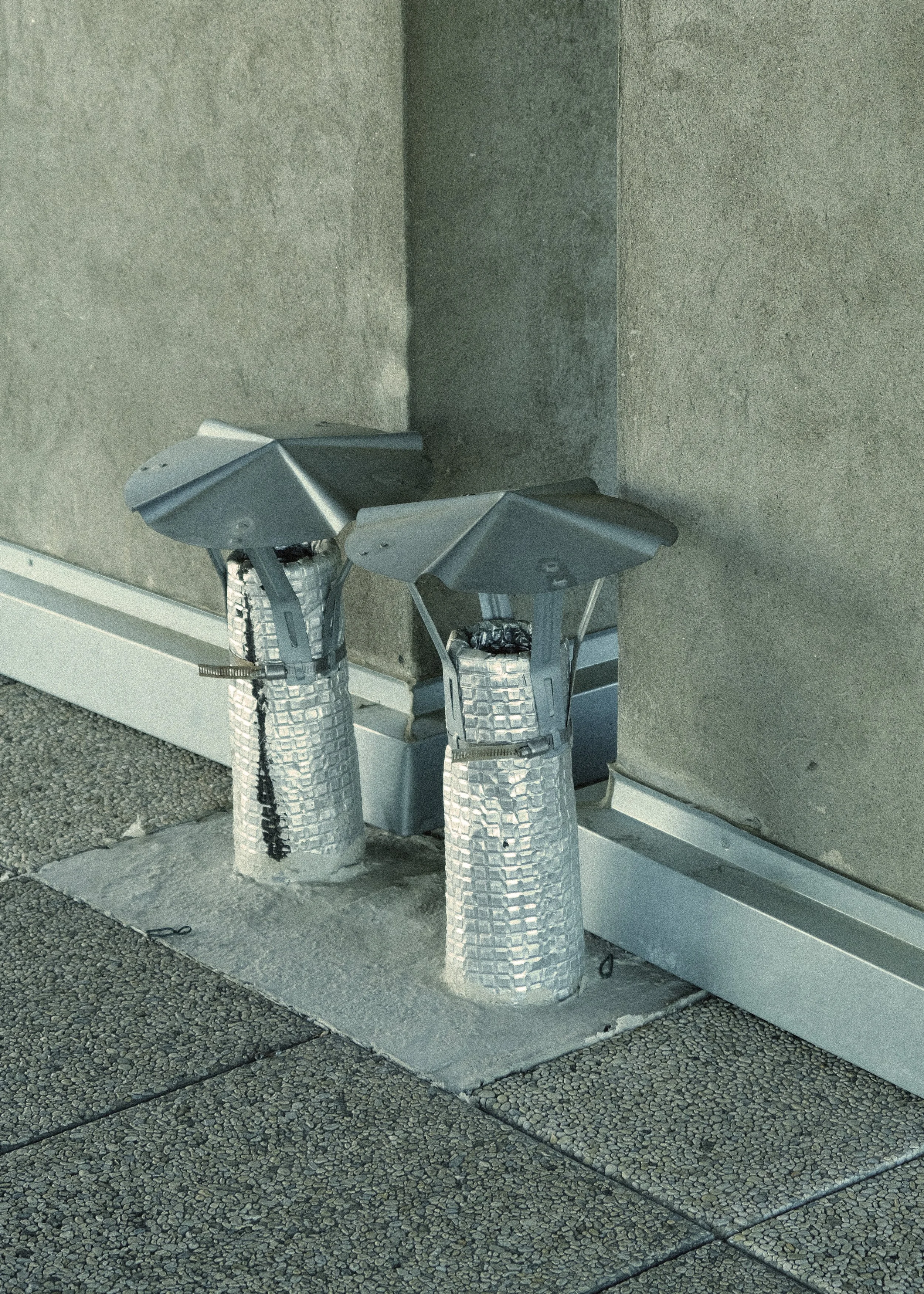 Trois purificateurs d'air en plastique blanc avec des filtres en tissu blanc, positionnés devant une paroi en béton gris et fixés au sol à l'aide de supports en métal.
