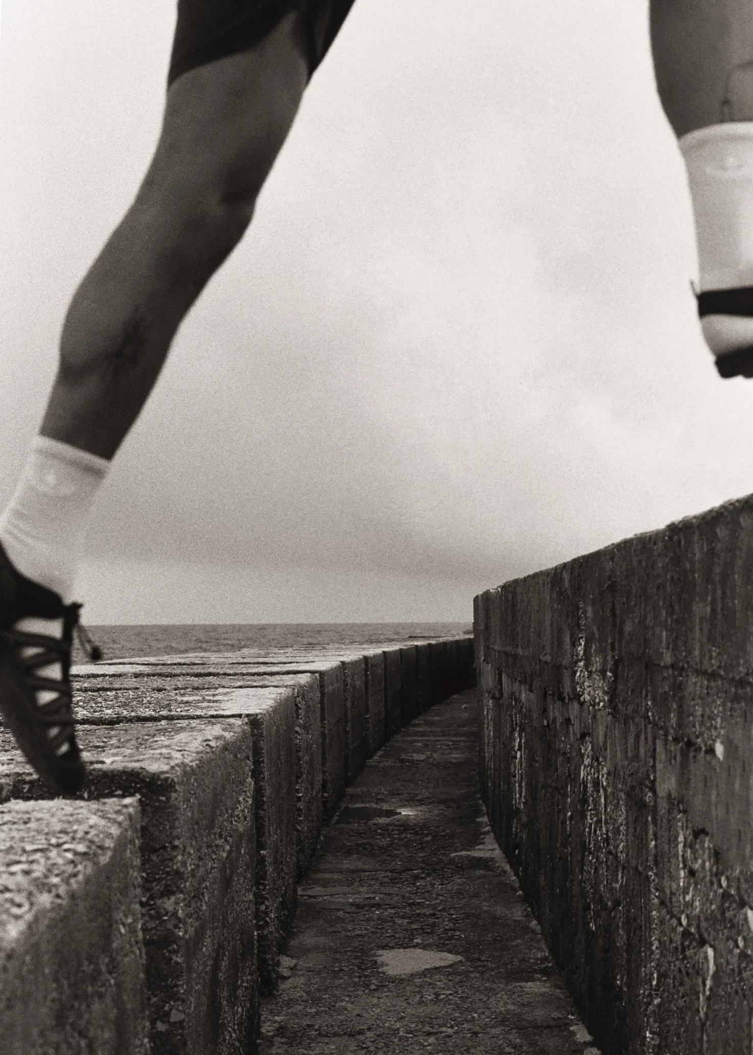 Photo en noir et blanc montrant une vue en contre-plongée entre deux murs en pierre, avec une personne qui marche à l'horizon, près de la mer ou de l'océan.