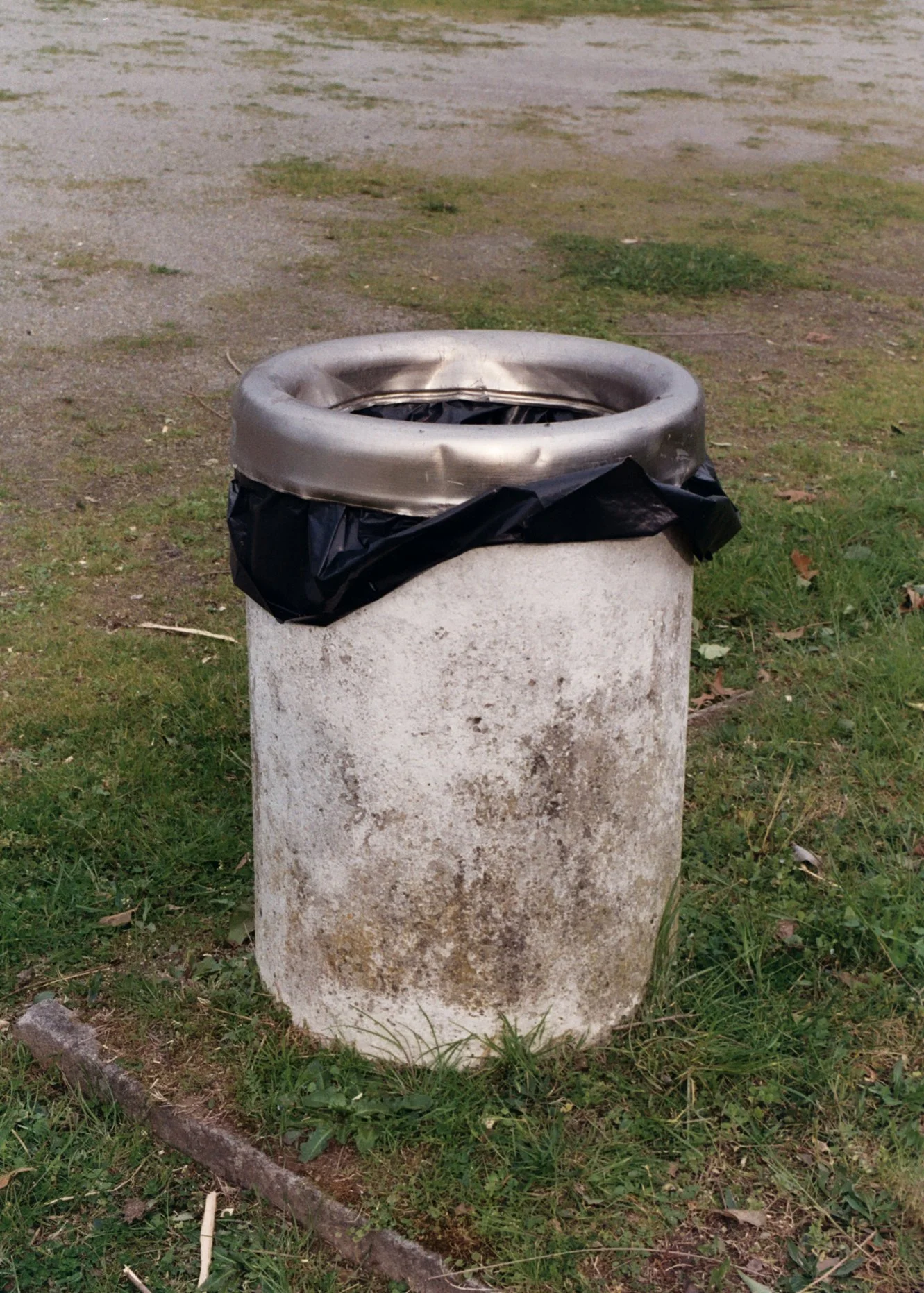 Poubelle en béton avec un sac en plastique noir à l'intérieur, située dans un espace vert avec de l'herbe et quelques branches au sol.