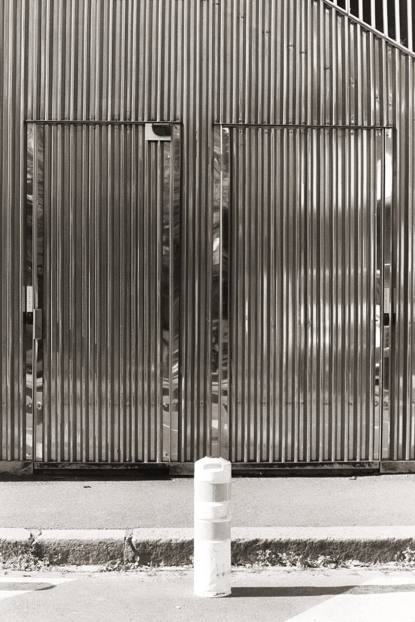 Une porte en métal à rainures verticales, un poteau en béton et une barrière de sécurité en plastique blanc sur le trottoir.