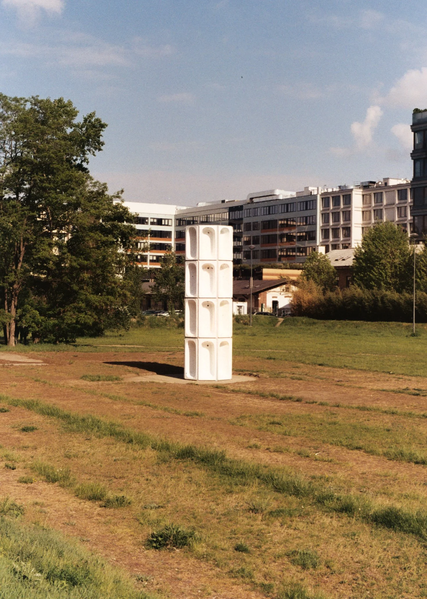 Un espace vert avec une sculpture blanche au centre, des bâtiments résidentiels en arrière-plan, des arbres et un ciel bleu avec quelques nuages.