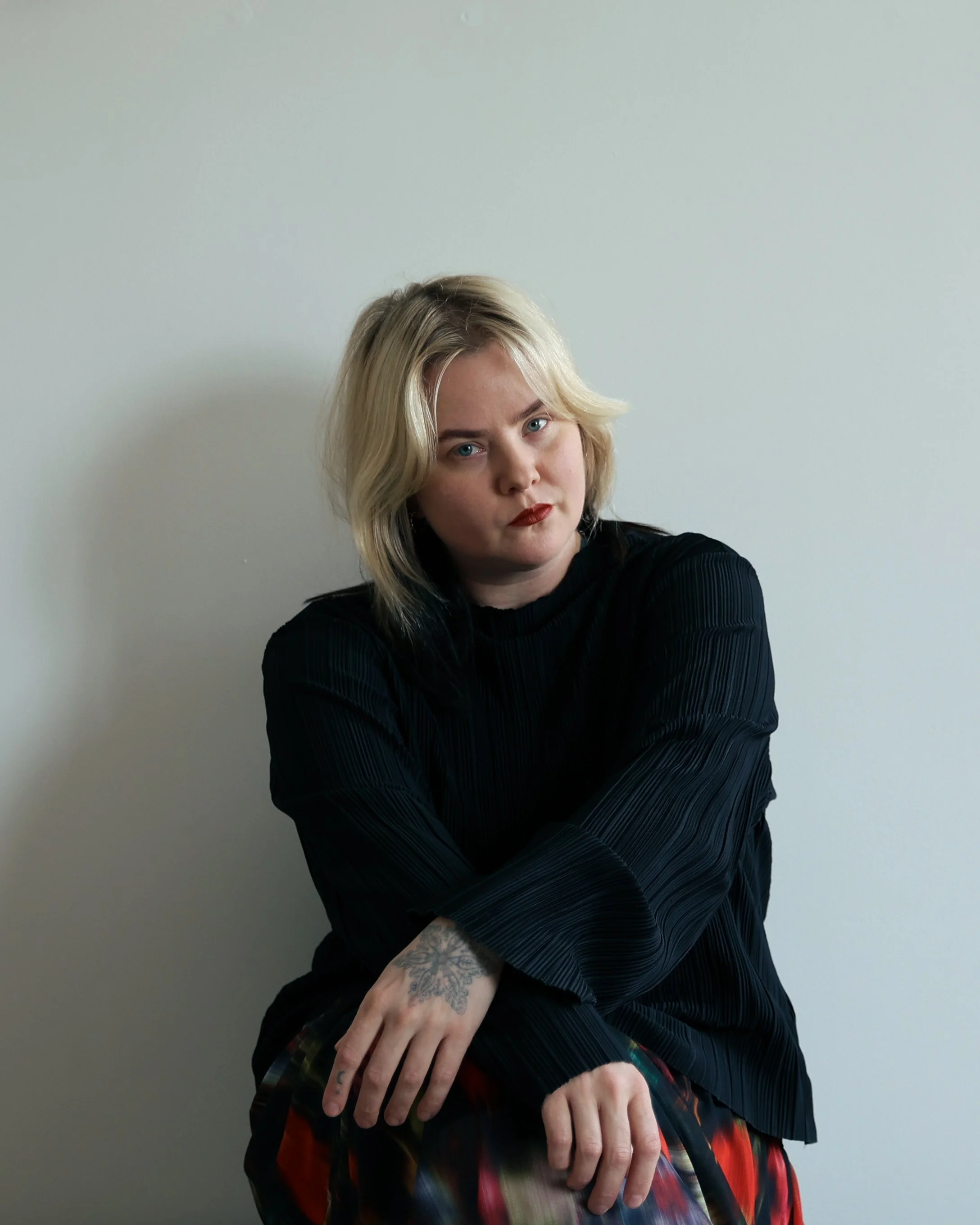 Une femme aux cheveux blonds, portant une veste noire à rayures fines, avec un maquillage rouge à lèvres, pose contre un mur blanc avec des bras croisés et une main visible avec tatouage.