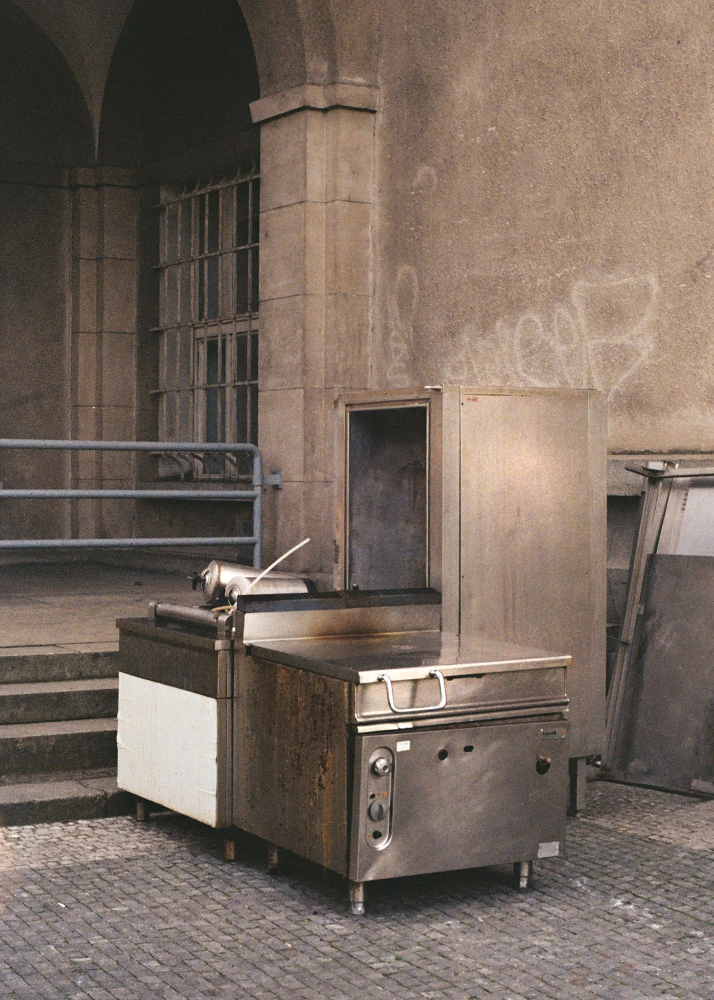 Le dos d'une machine de cuisson en acier inoxydable sur un trottoir pavé dehors, avec un garage ou une ancienne bâtisse en pierre en arrière-plan.