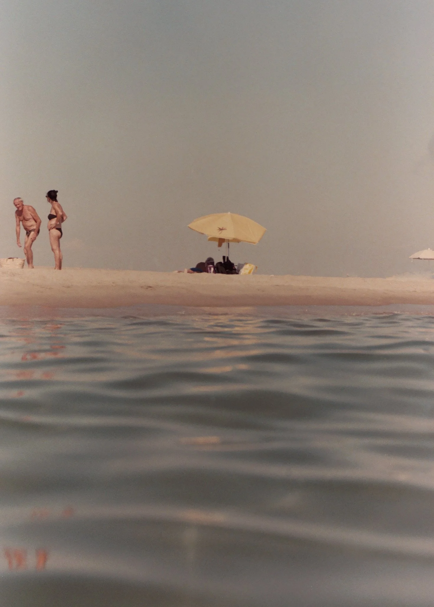 Vue de la plage avec deux personnes en maillot de bain, une ombrelle jaune, et un ciel clair.